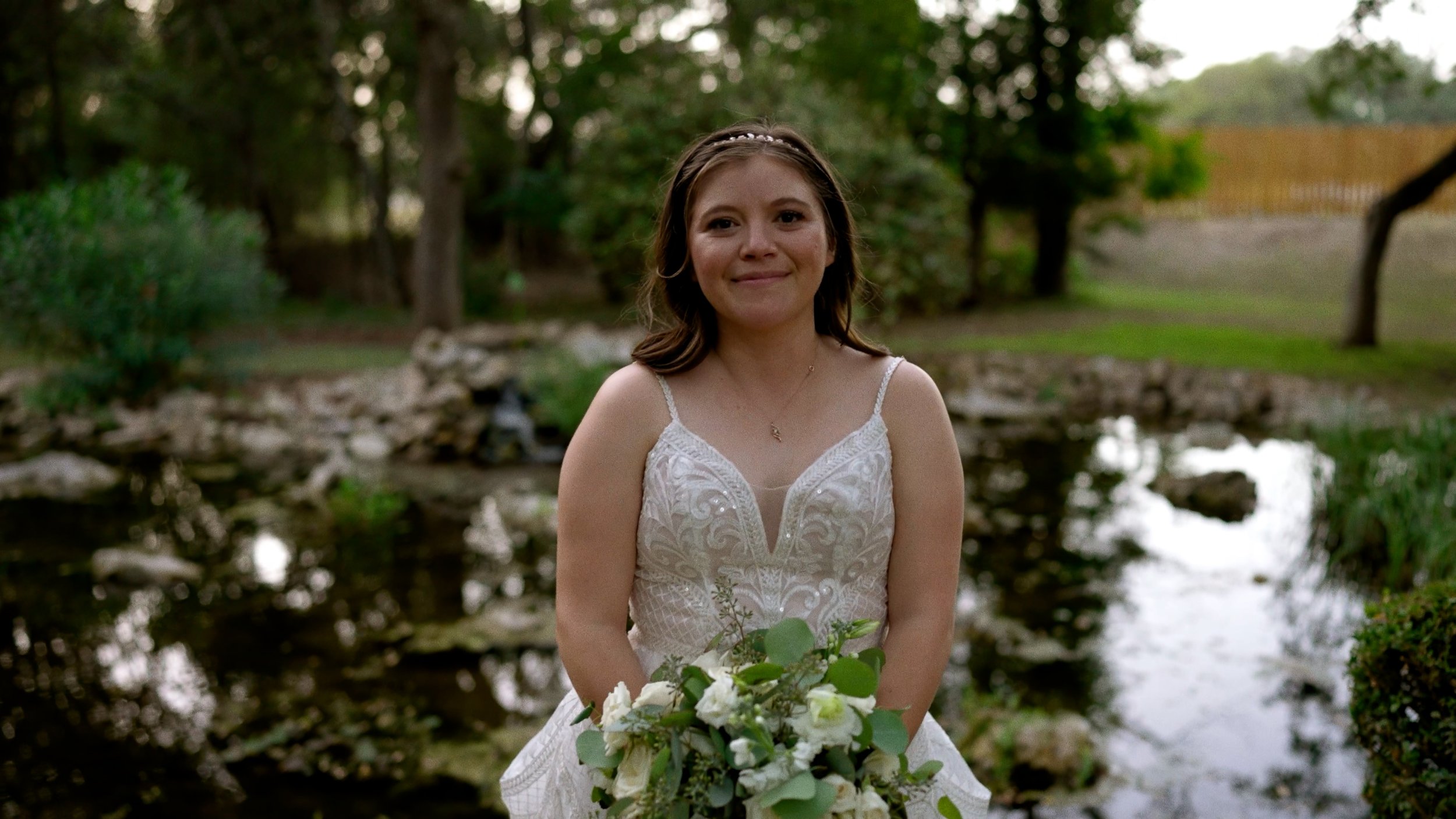 bride soft smiles at camera holding bouquet in wedding dress outside next to pond