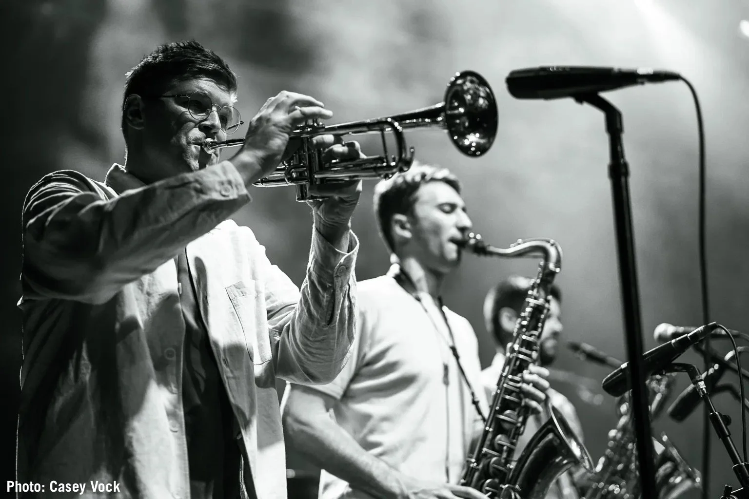 Black and white photo of three musicians playing instruments on stage. The first musician, a man with glasses, is playing a trumpet. Two other musicians are playing saxophones in the background.
