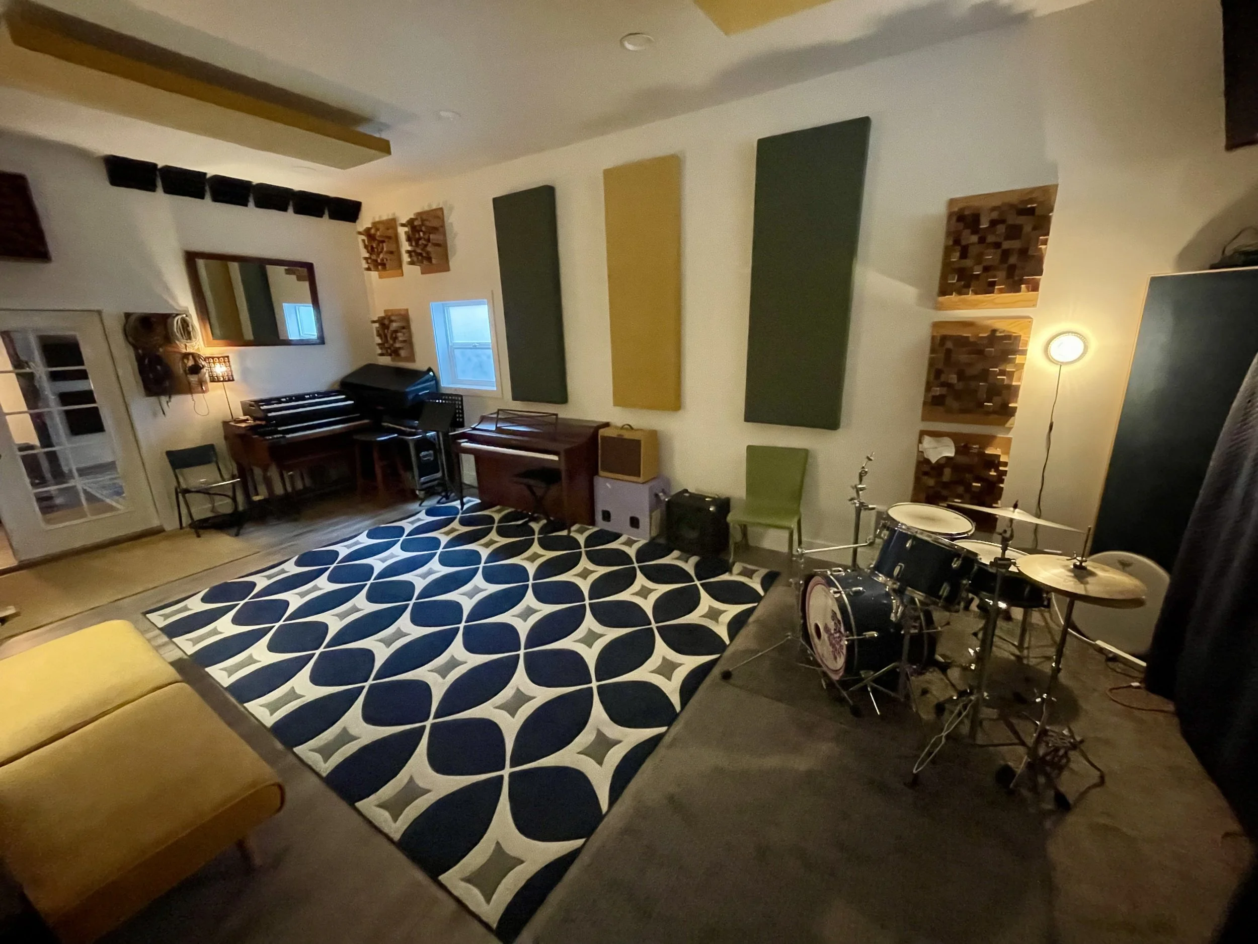 A music studio with acoustic panels on the wall, a window, a mirror, a keyboard, a small amplifier, a green chair, a drum set, and a patterned rug.