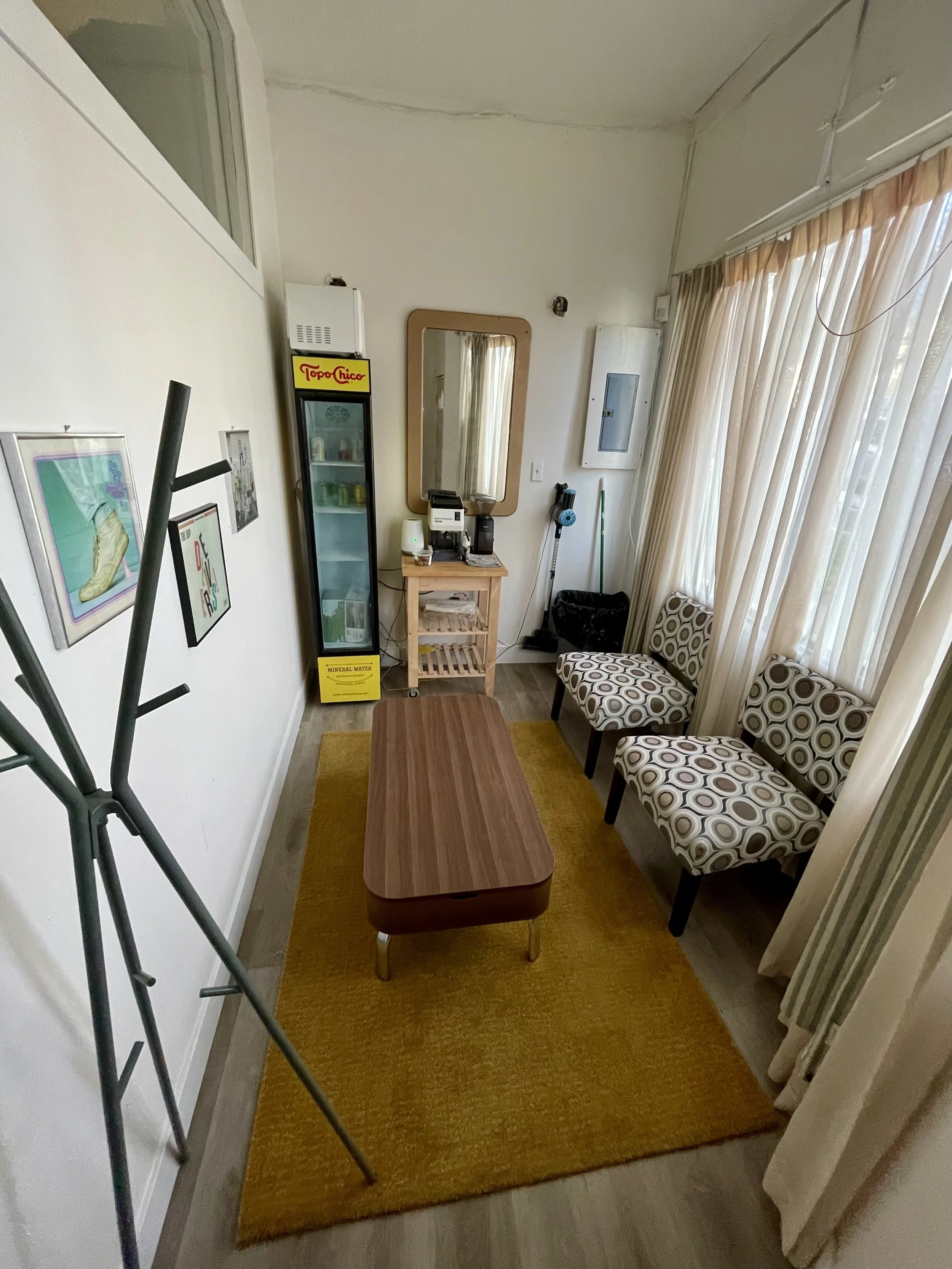 Small living room with a mustard yellow rug, a wooden coffee table, a booth with patterned upholstery, a mirror, shelves, and curtains covering a window.