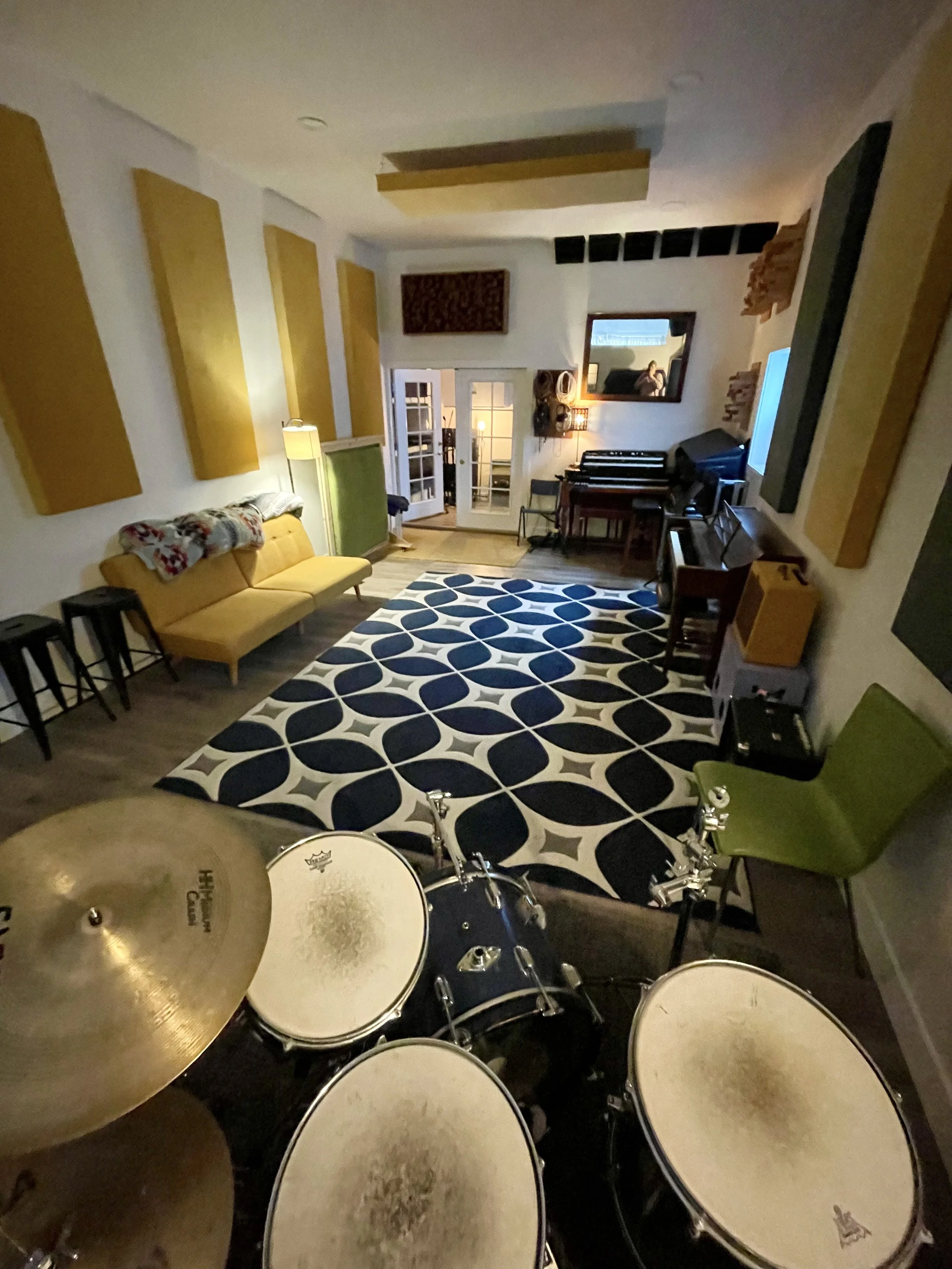 A living room with a yellow couch, a blue and white patterned rug, a drum set, and musical instruments like a keyboard and an amplifier, with a mirror reflecting a person taking the photo.
