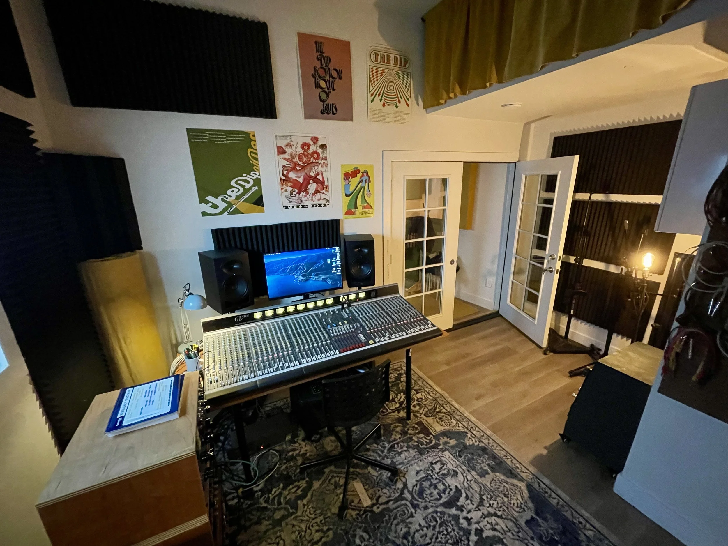 A recording studio with soundproofed walls, posters on the wall, a mixing console, a computer monitor, a desk with books, chairs, and a patterned rug on the floor.