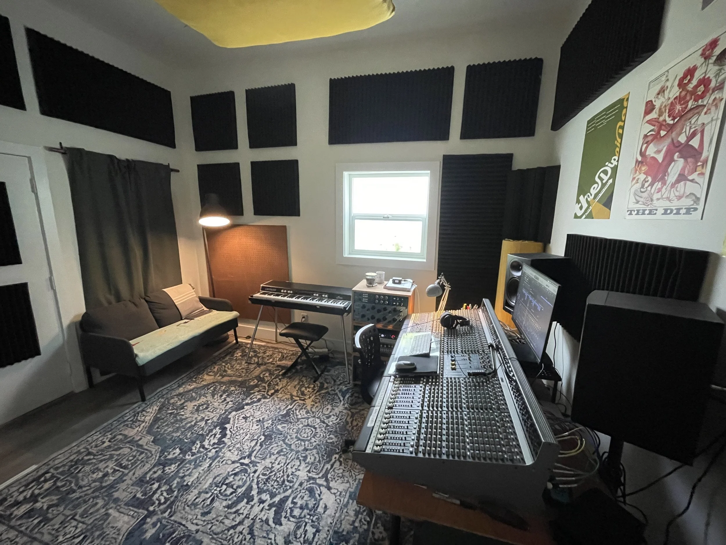 A music recording studio with soundproofing foam on the walls, a window, a couch, and musical equipment including a keyboard, mixing console, and computer monitor, all on an ornate rug.