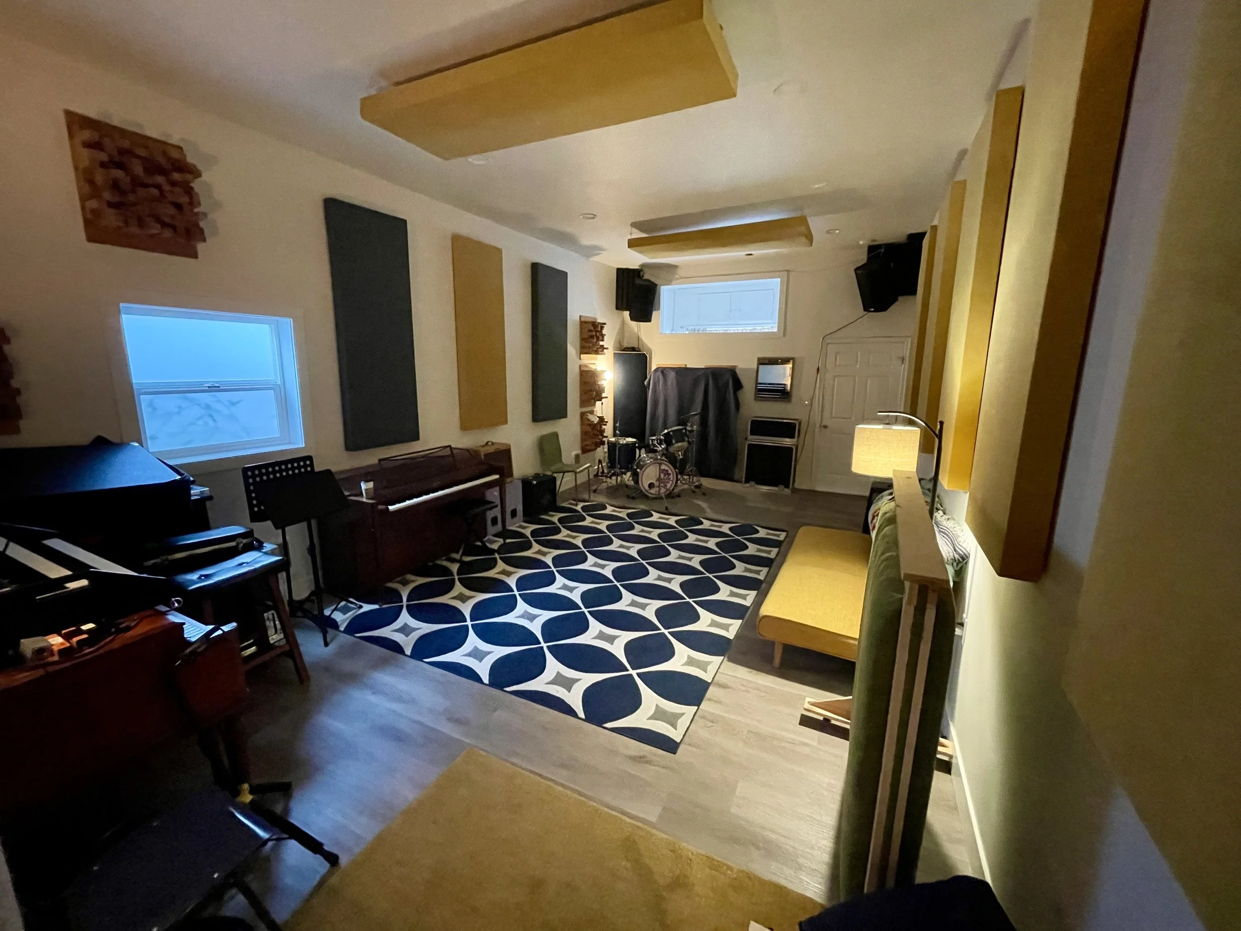 Living room with two windows, acoustic wall panels, a patterned rug, a drum set, a piano, and a yellow bench.