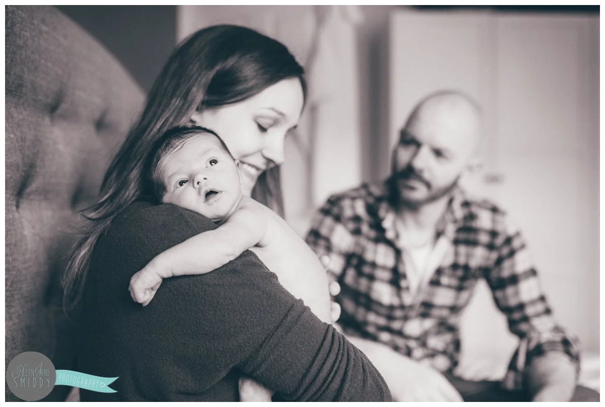 Beautiful black and white photograph of a newborn baby girl being held by her mother with her father in the background.
