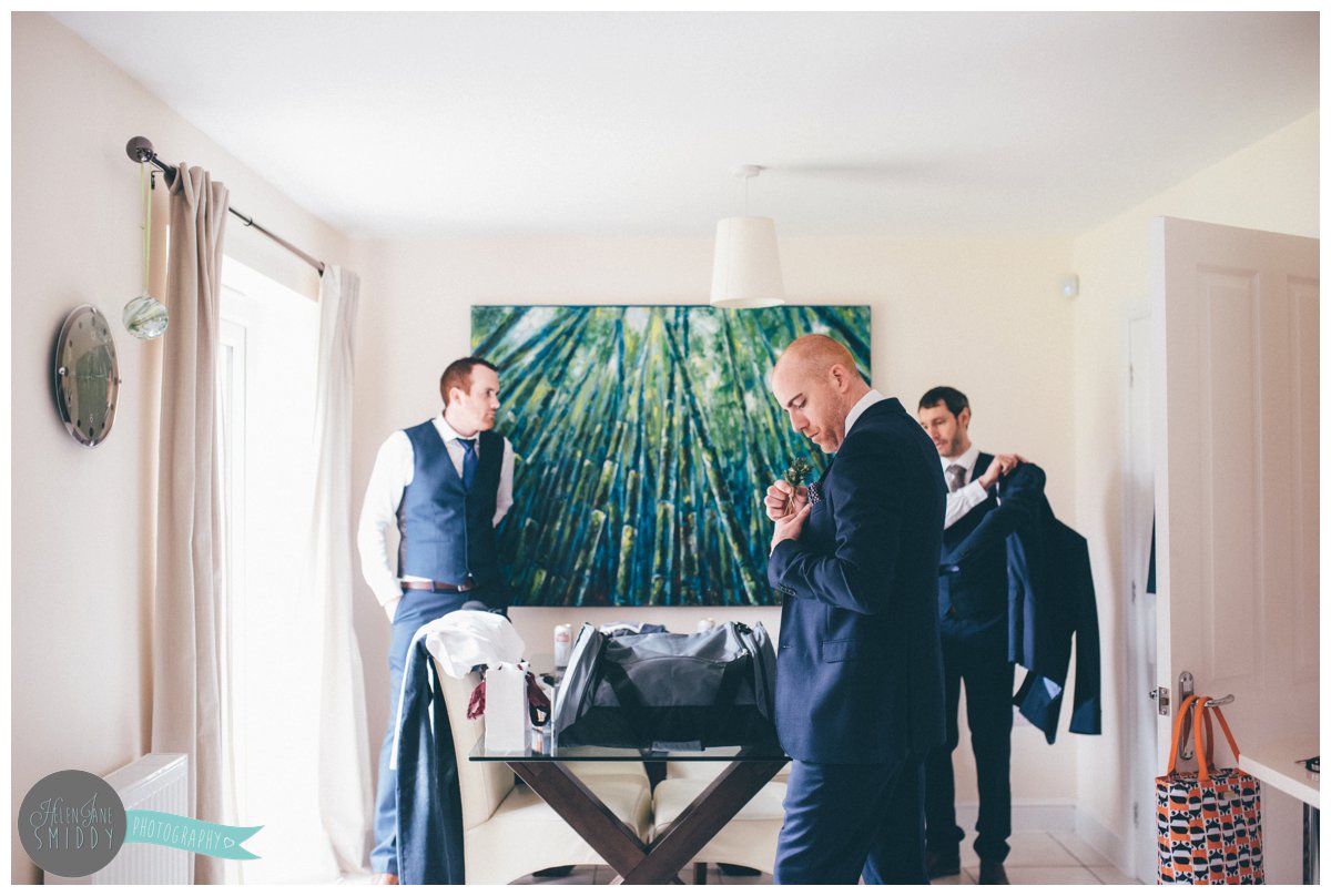 The groom arranges his hanky as his Best Man puts his suit jacket on and his usher smiles.