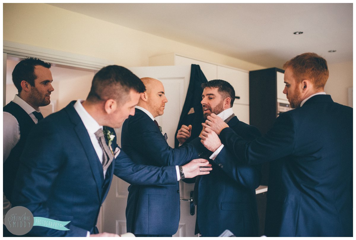 The groom and all the groomsmen all help their one friend tie his tie and sort out his suit.