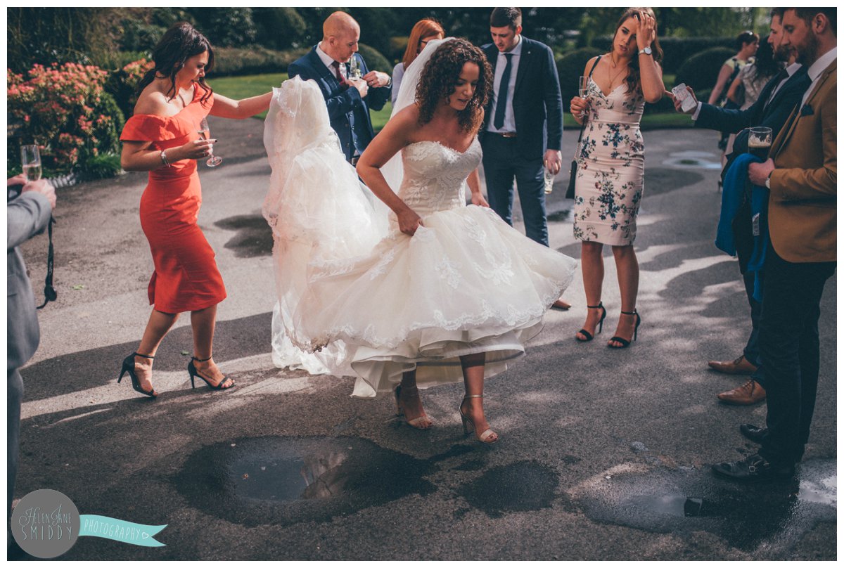 Lady in reed helps the bride protect her wedding gown from the puddles whilst the walk into Mer Court Hotel.