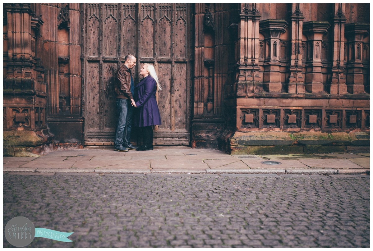 Couple face each other and hold hands, whilst stood in front of the beautiful big brown door of Chester Cathedral during their engagement shoot.