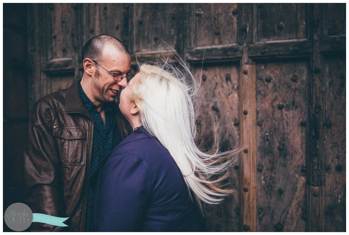 Close-up of couple facing each other and hold hands, whilst stood in front of the beautiful big brown door of Chester Cathedral during their engagement shoot.