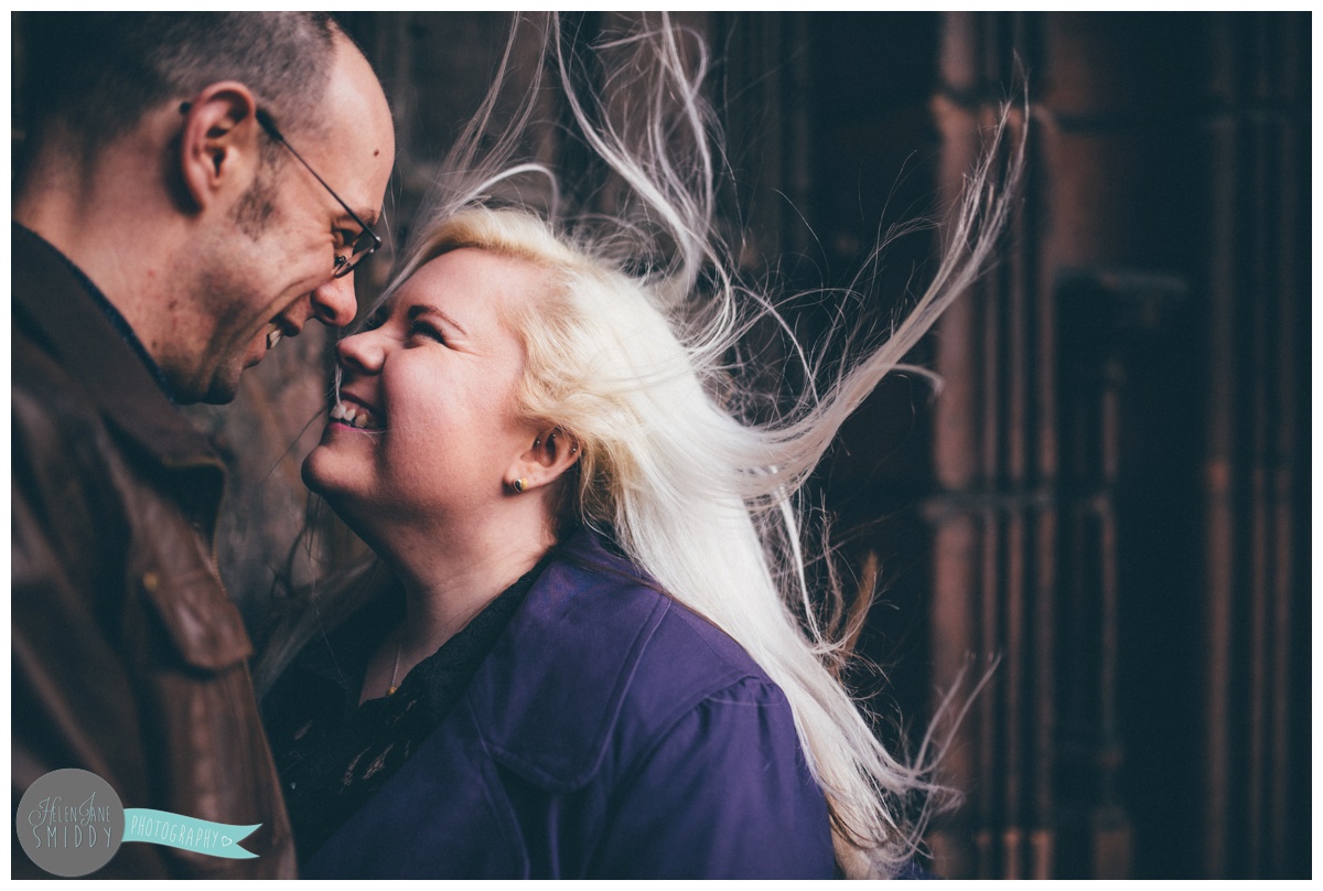 The wind blows the girl's hair as the couple face each other and hold hands, whilst stood in front of the beautiful big brown door of Chester Cathedral during their engagement shoot.