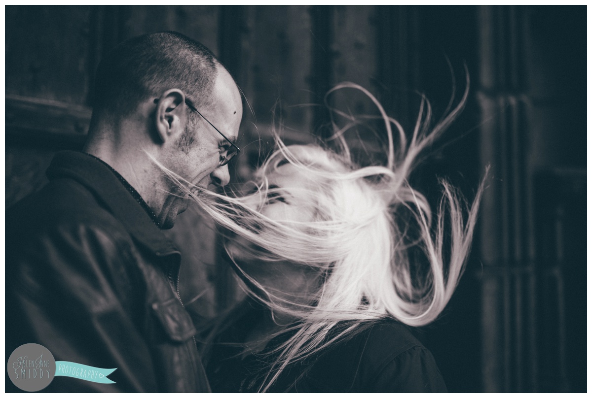 The wind covers the bride's face with her hair as the couple face each other and hold hands, whilst stood in front of the beautiful big brown door of Chester Cathedral during their engagement shoot.