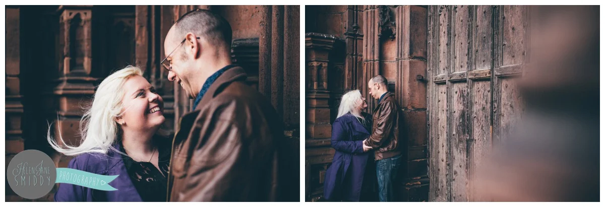 Couple face each other and hold hands, whilst stood in front of the beautiful big brown door of Chester Cathedral during their engagement shoot.