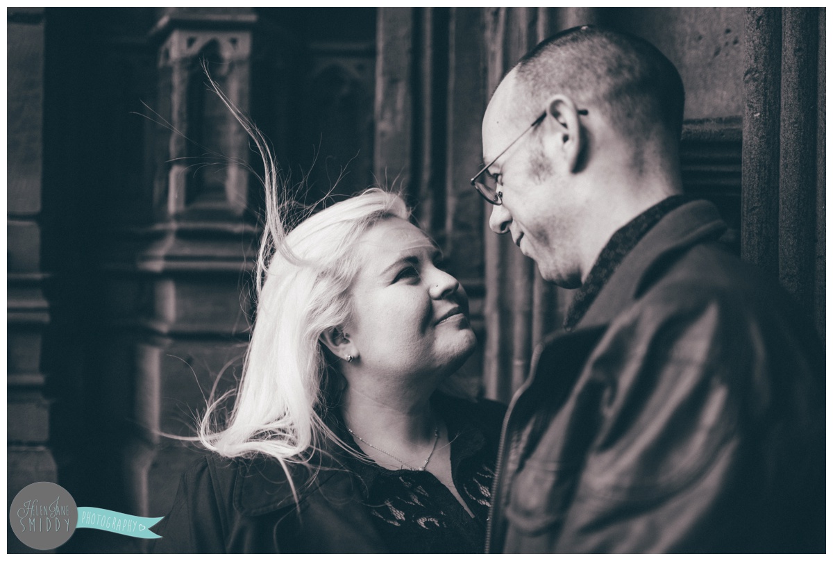 Couple face each other and hold hands, whilst stood in front of the beautiful big brown door of Chester Cathedral during their engagement shoot in Chester City Centre.