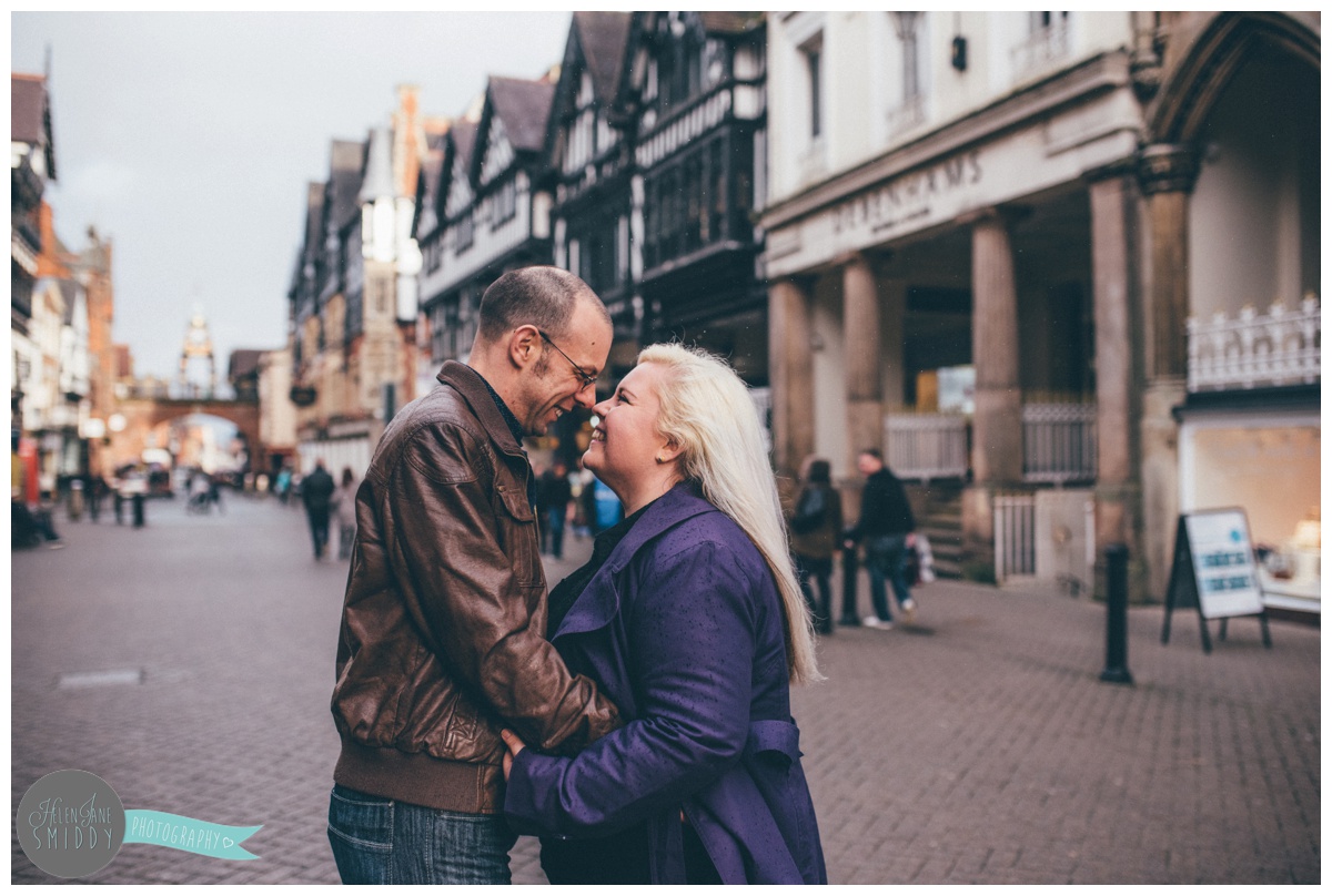 Jo and Alan stand, hugging and laughing in Chester City Centre for their engagement shoot.