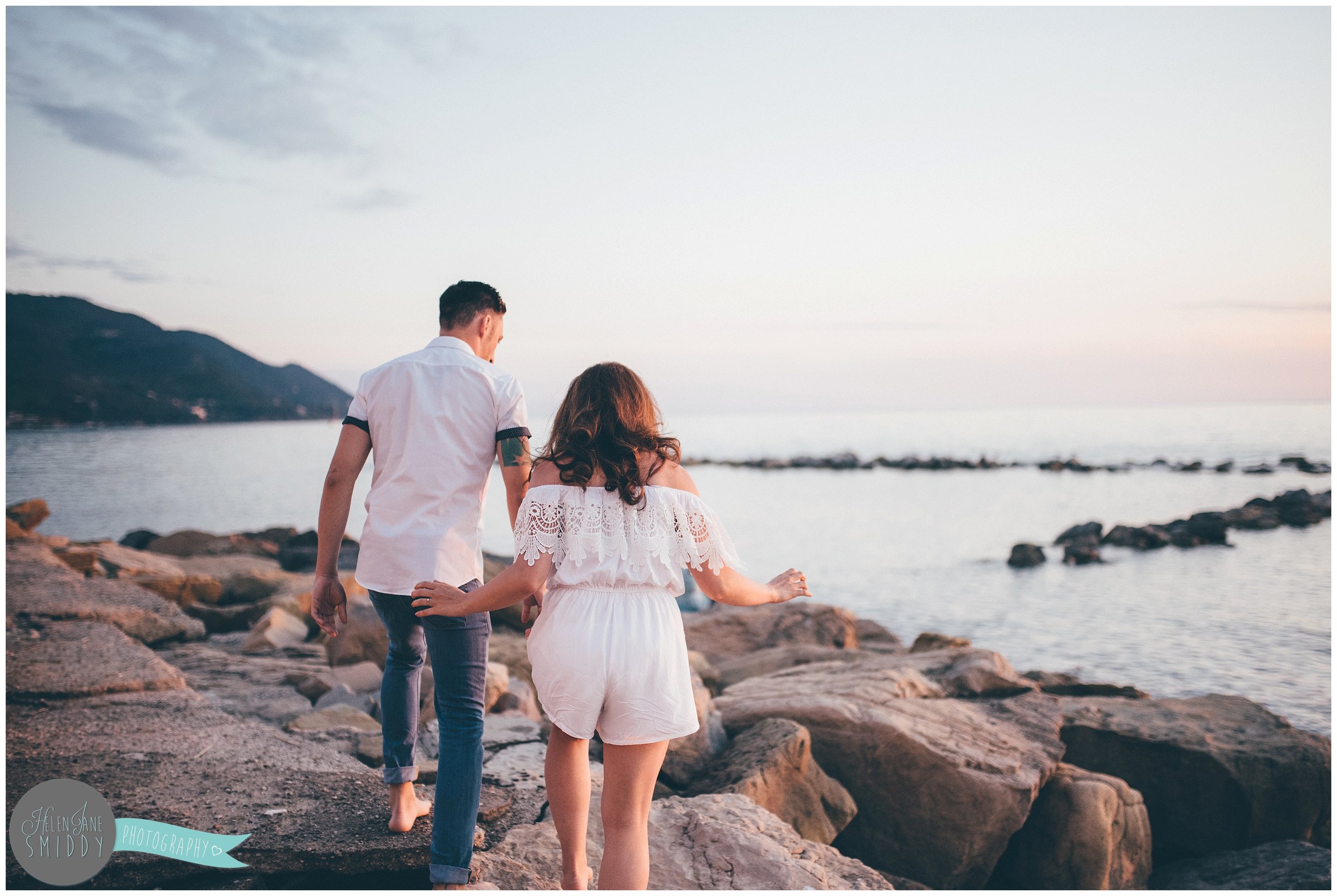 The young couple climb rocks on the beach during their Engagement photoshoot in Santa Maria Di Castellabate, Italy.