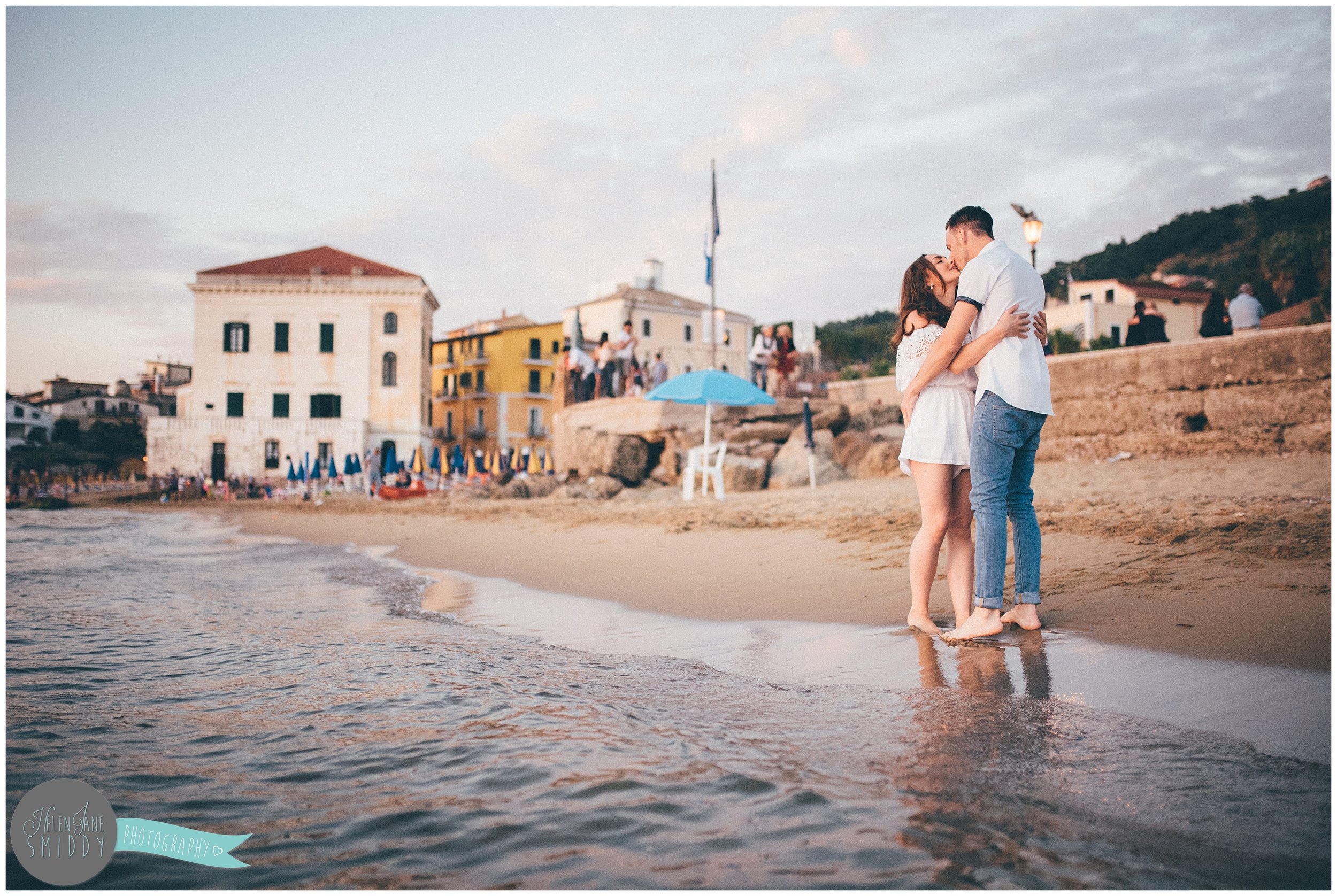 The couple stand in the water kissing, with Santa Maria di Castellabate in the background.