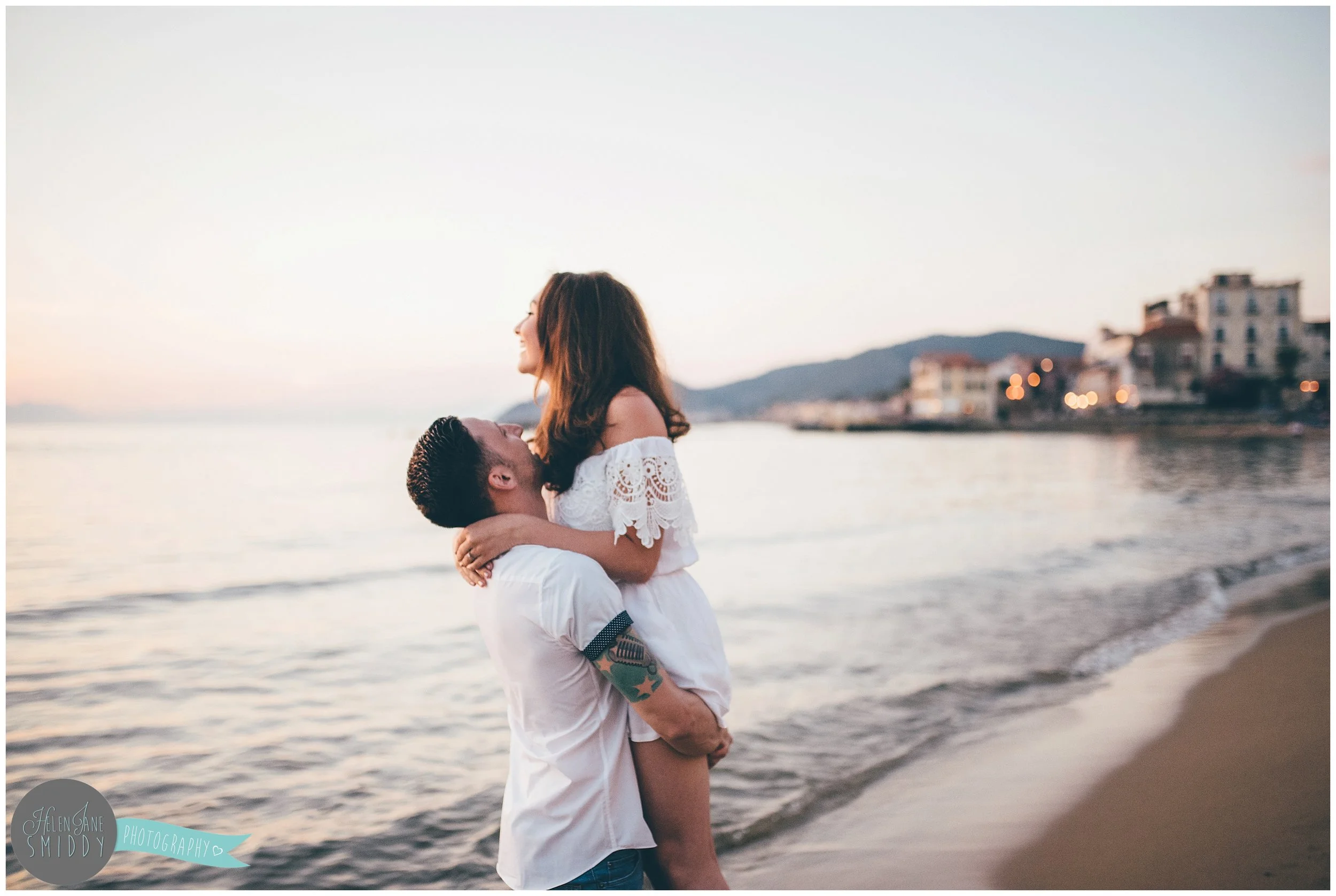 Dom lifts up his beautiful fiancee on the beach at sunset.