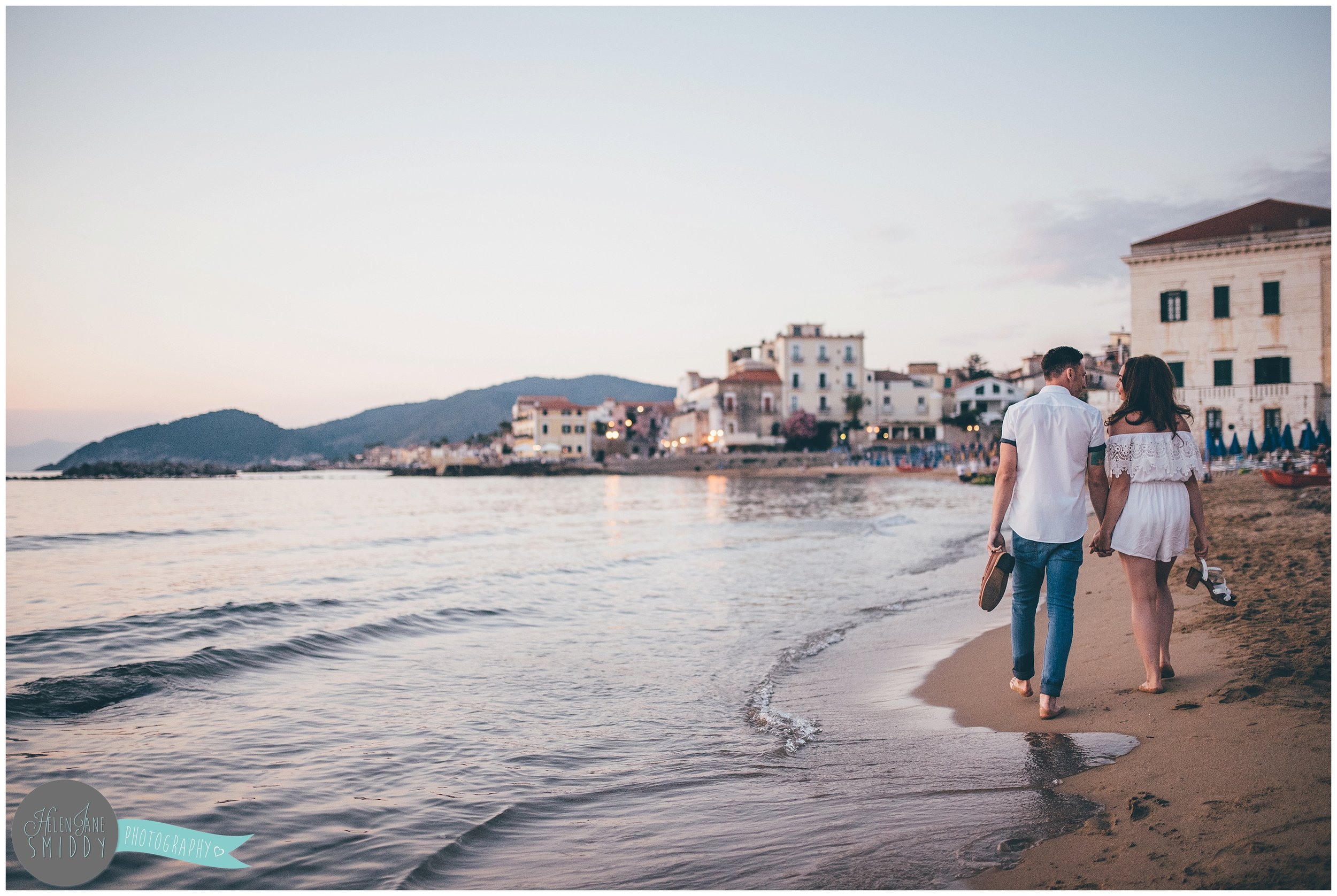 The couple walk hand-in-hand across the beach with Santa Maria di Castellabate in the background.