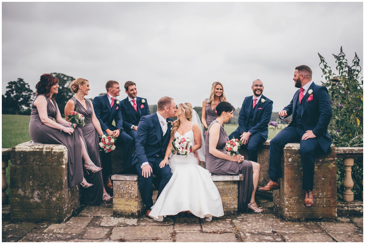 The bride and groom share a kiss whilst the bridal party sit around them chatting in the pretty gardens at Willington Hall.