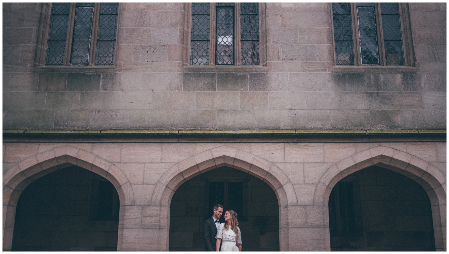 Bride and groom stand together outside gothic old English building in Manchester.