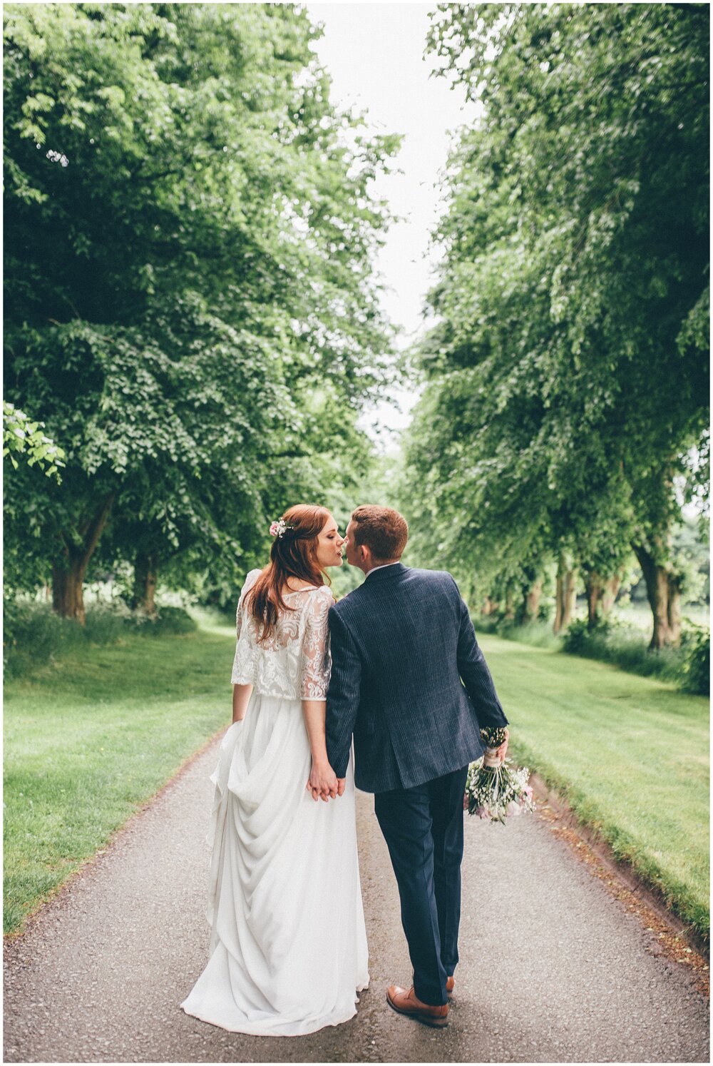 Bride and groom stroll hand in hand down a tree-lined path in Cheshire.
