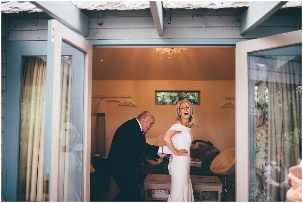 Bride laughs as her dad fastens her into her beautiful Suzanne Neville wedding gown.