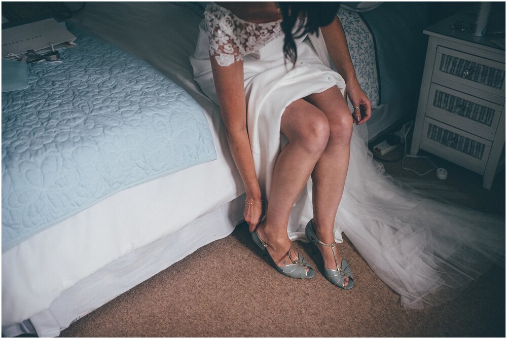 Bride sits on the bed and puts on her Rachel Simpson wedding shoes.