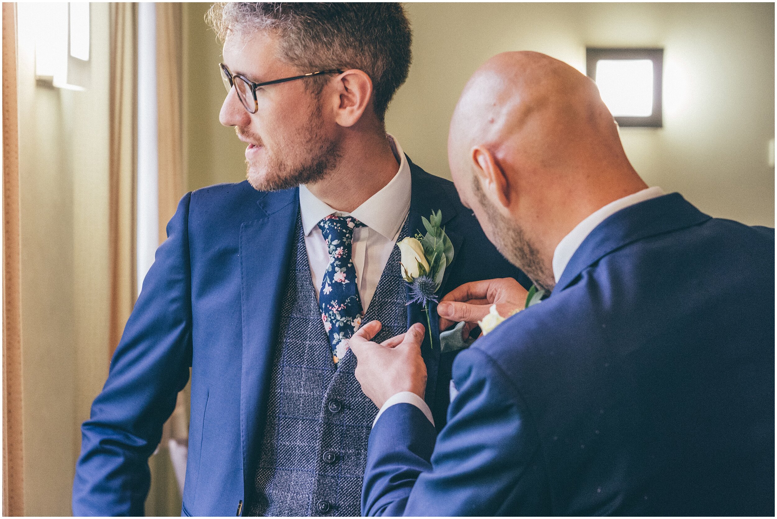 Best Man helps the groom fasten on his button hole at Quarry Bank Mill wedding in Cheshire near to Manchester.