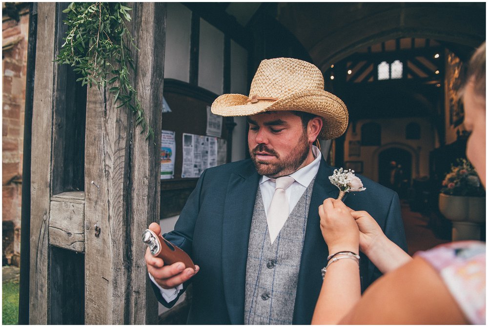 Groomsman wears a cowboy hat and drinks from his hip flask as someone fastens his buttonhole on