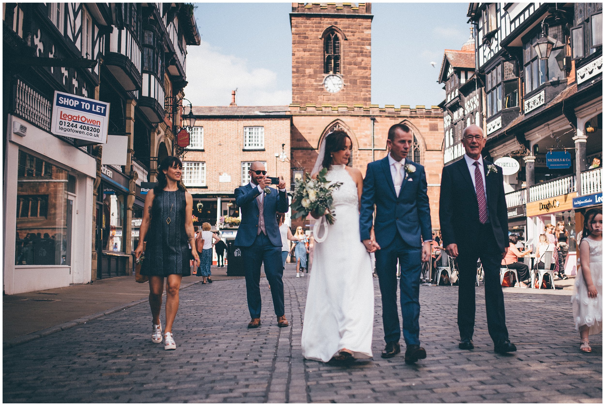 Guest takes a photograph of bride and groom walking through Chester city centre