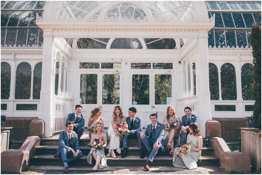 Bridesmaids and groomsmen sit with the bride and groom on the steps of Sefton Palm House in Liverpool.