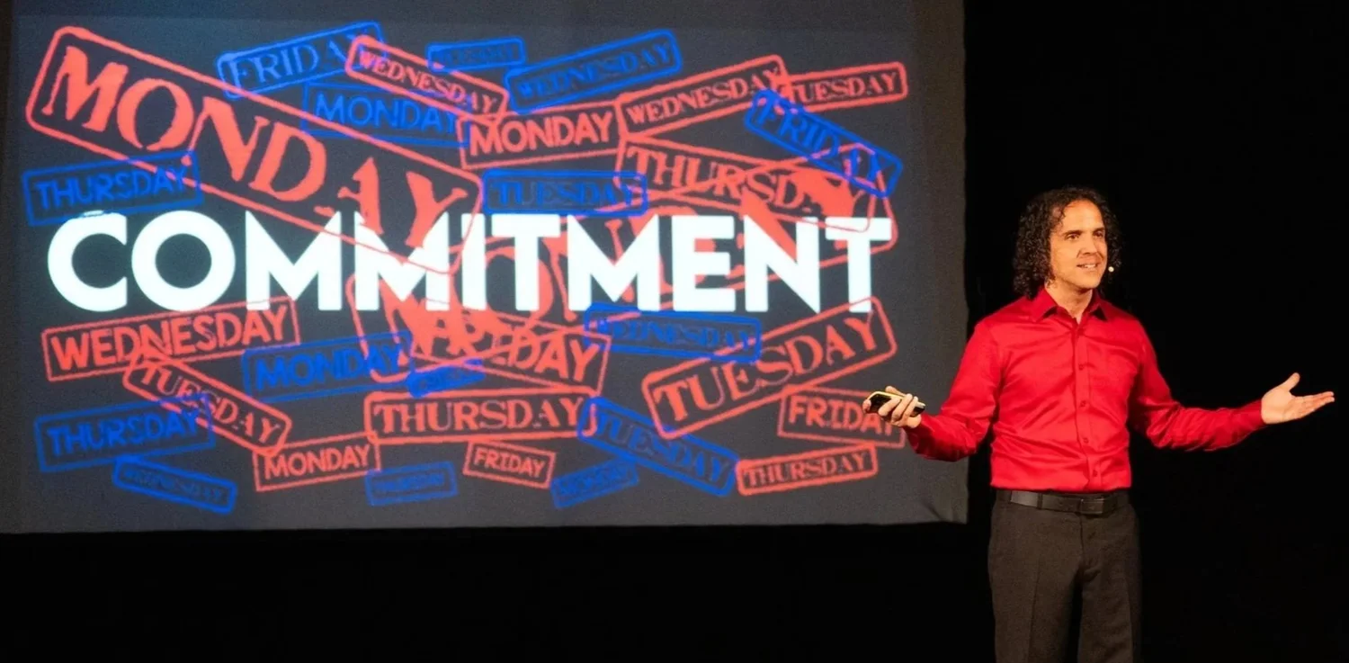 Davide Di Giorgio on stage in a red shirt, in front of a screen filled with Monday to Friday stamps and the word Commitment