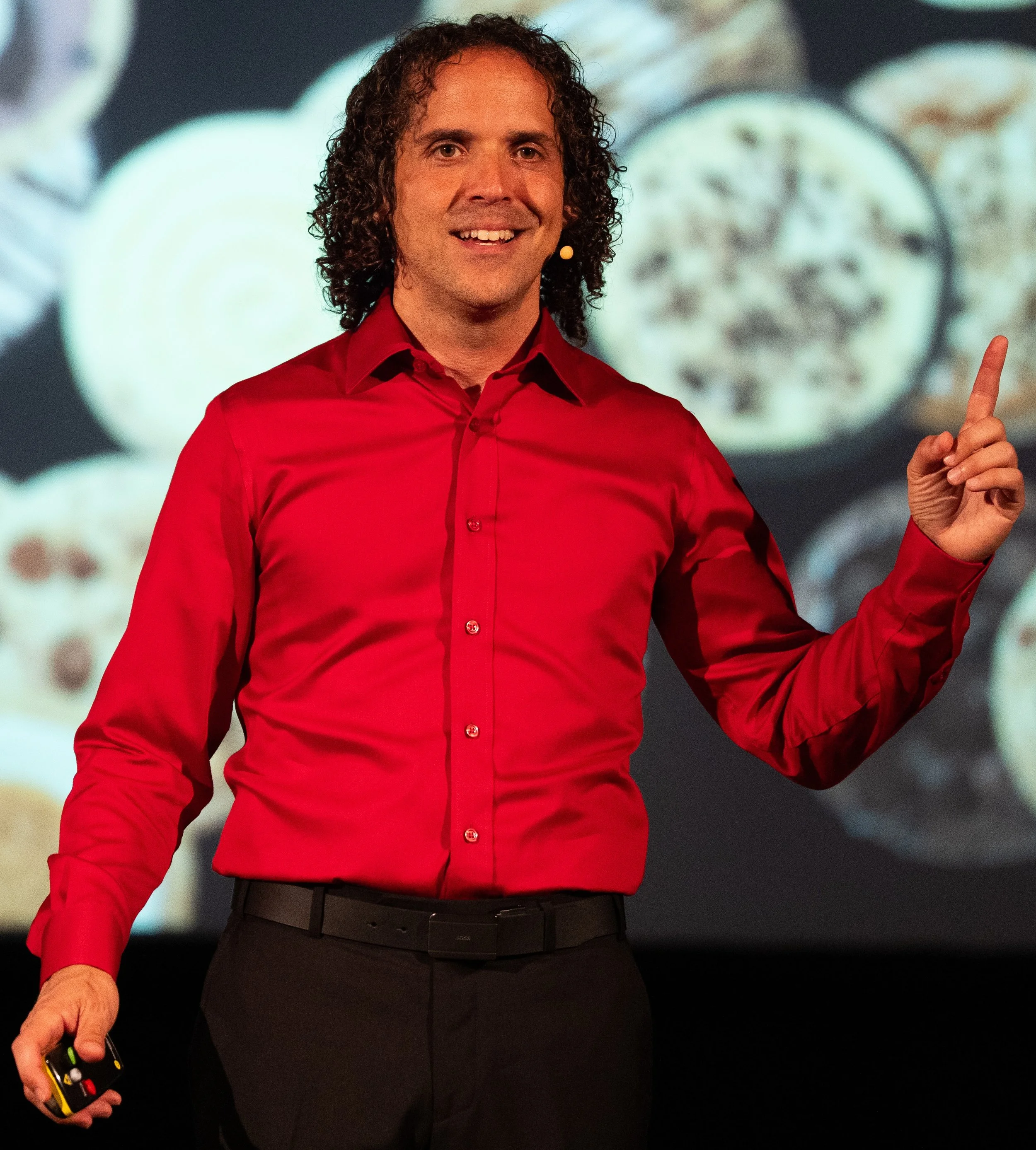 Davide Di Giorgio, Operations & Leadership Advisor and Keynote Speaker, on stage mid-keynote in a red shirt, one finger raised to make a point, with a large projected image of cookies behind him.