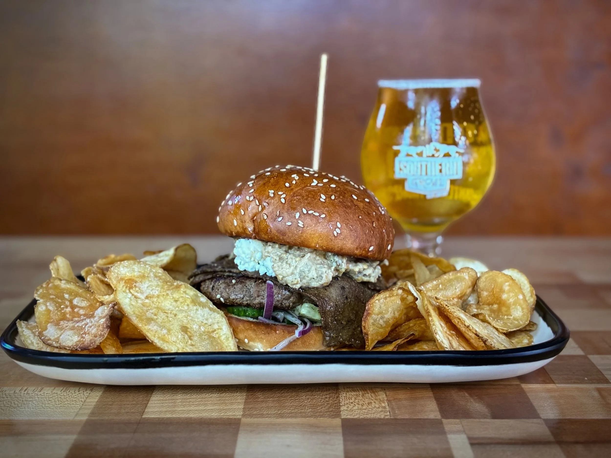 FEAST FROM THE EAST BURGER topped with spicy baba ganoush, shaved lamb, cucumber salad, crumbled feta, and za&rsquo;atar on a house sesame bun. Special available through Sunday during lunch, dinner, and brunch. 
Pair with GREER BEER, premium lager. A