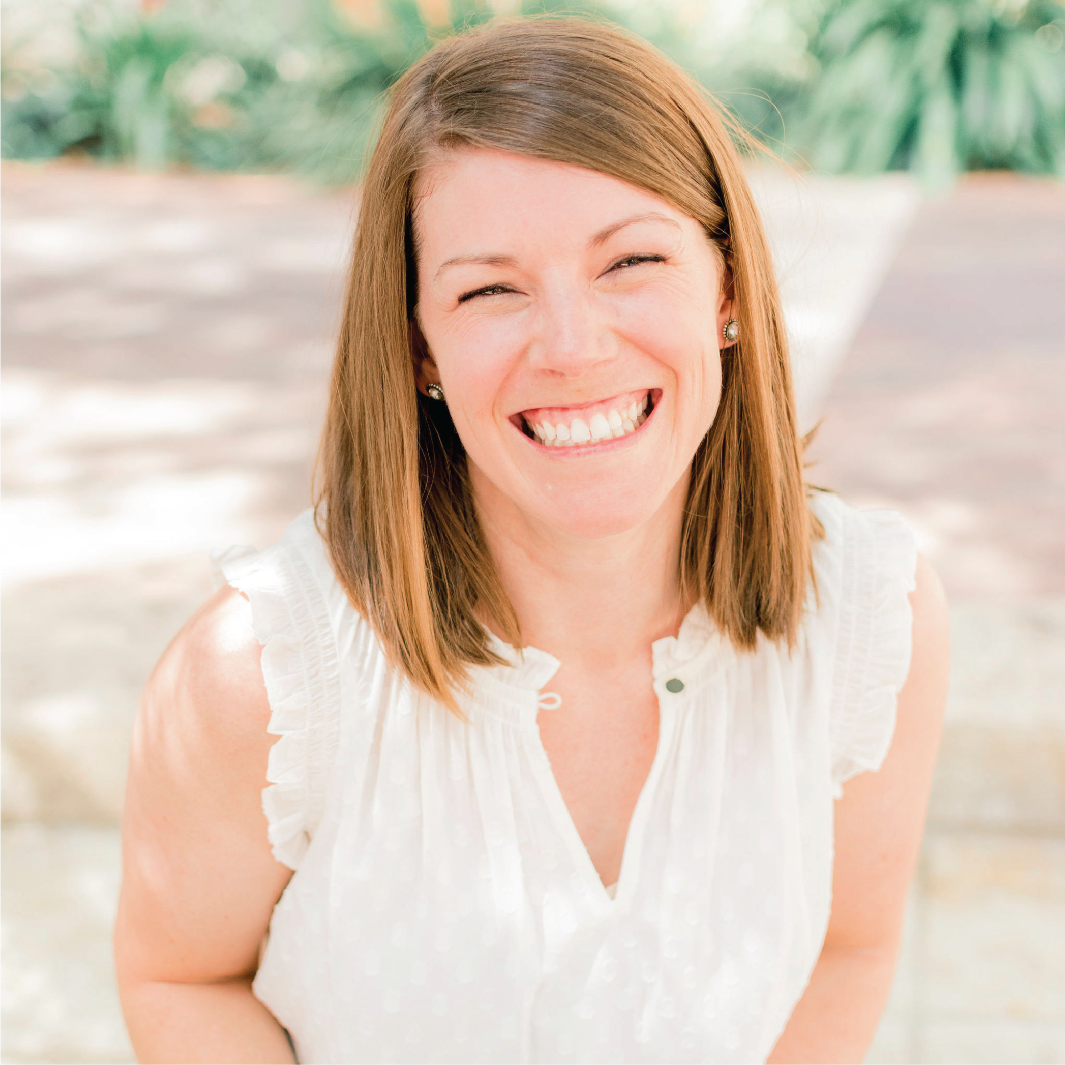 A woman with shoulder-length brown hair smiling and showing her teeth, wearing a sleeveless white top with ruffled shoulders, outdoors with blurred greenery in the background.