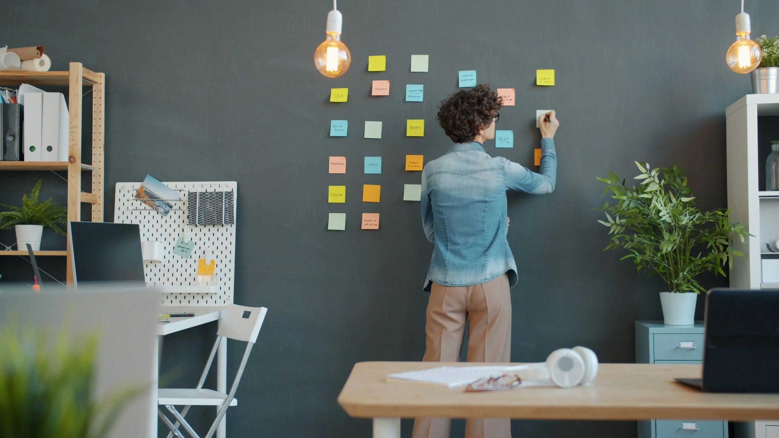 A person with curly hair and glasses writing on sticky notes on a wall covered with other sticky notes in an office or workspace. The room has a chalkboard wall, shelves with books and plants, and a table with headphones and papers.