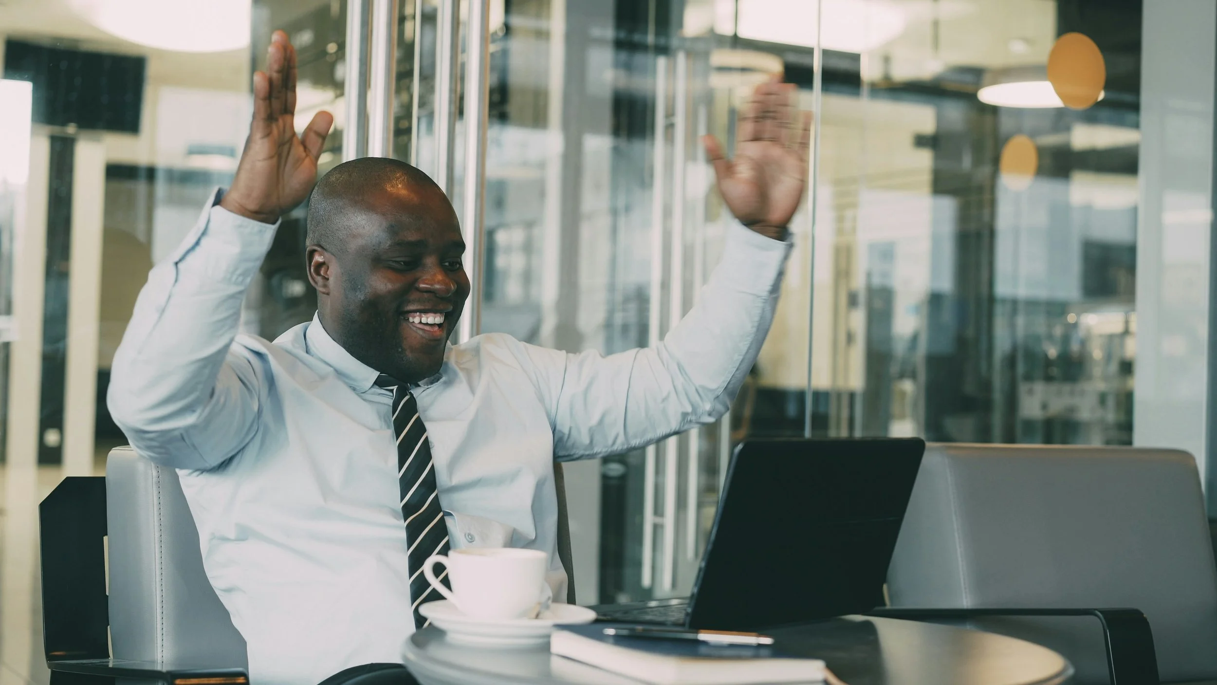 A man in a white shirt and striped tie smiling and raising his hands while sitting at a table in a modern office cafe, with a laptop, a cup of coffee, and a notebook in front of him.