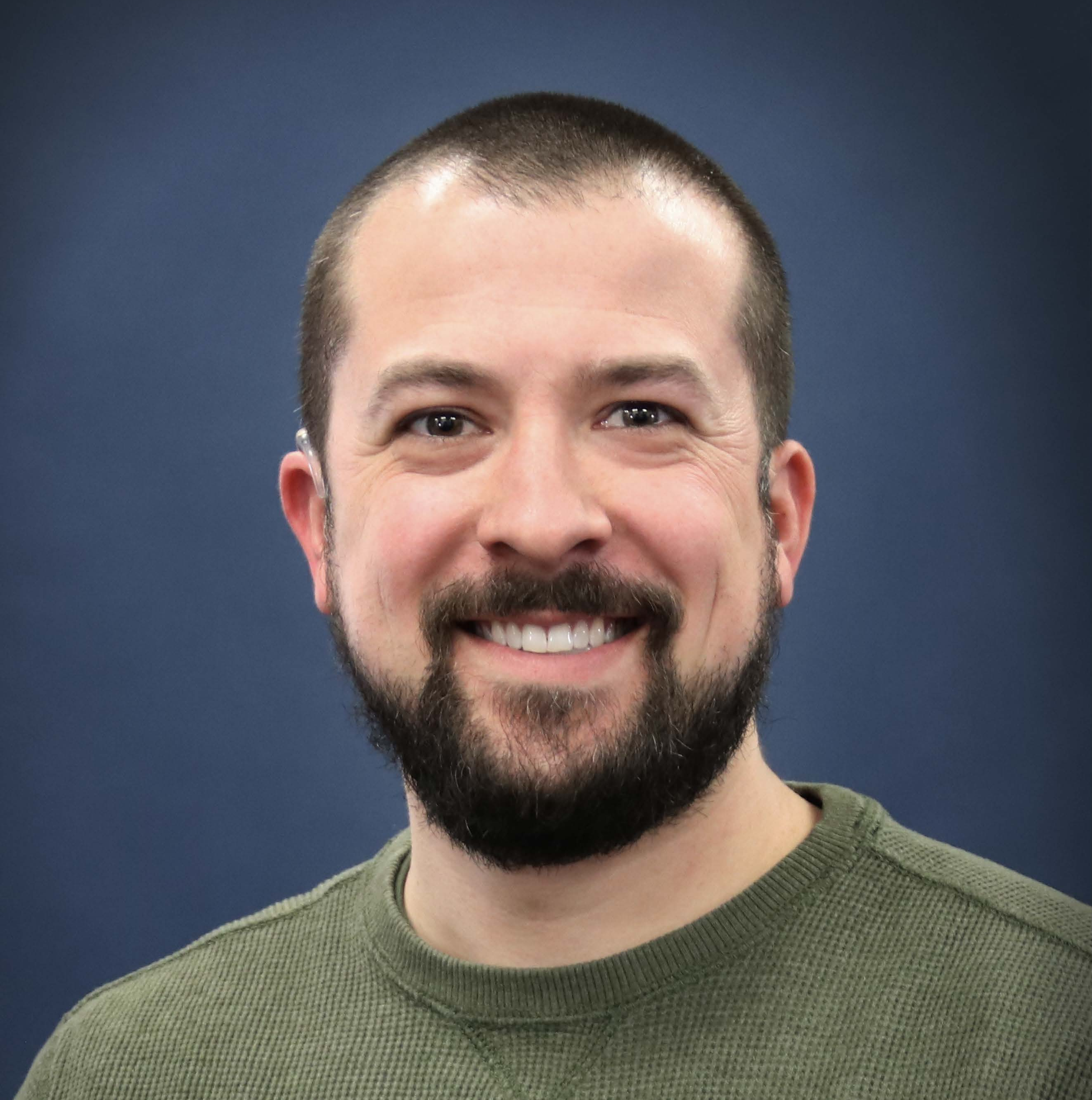 A smiling man with short brown hair, a beard, wearing a green sweater, standing against a solid dark blue background.