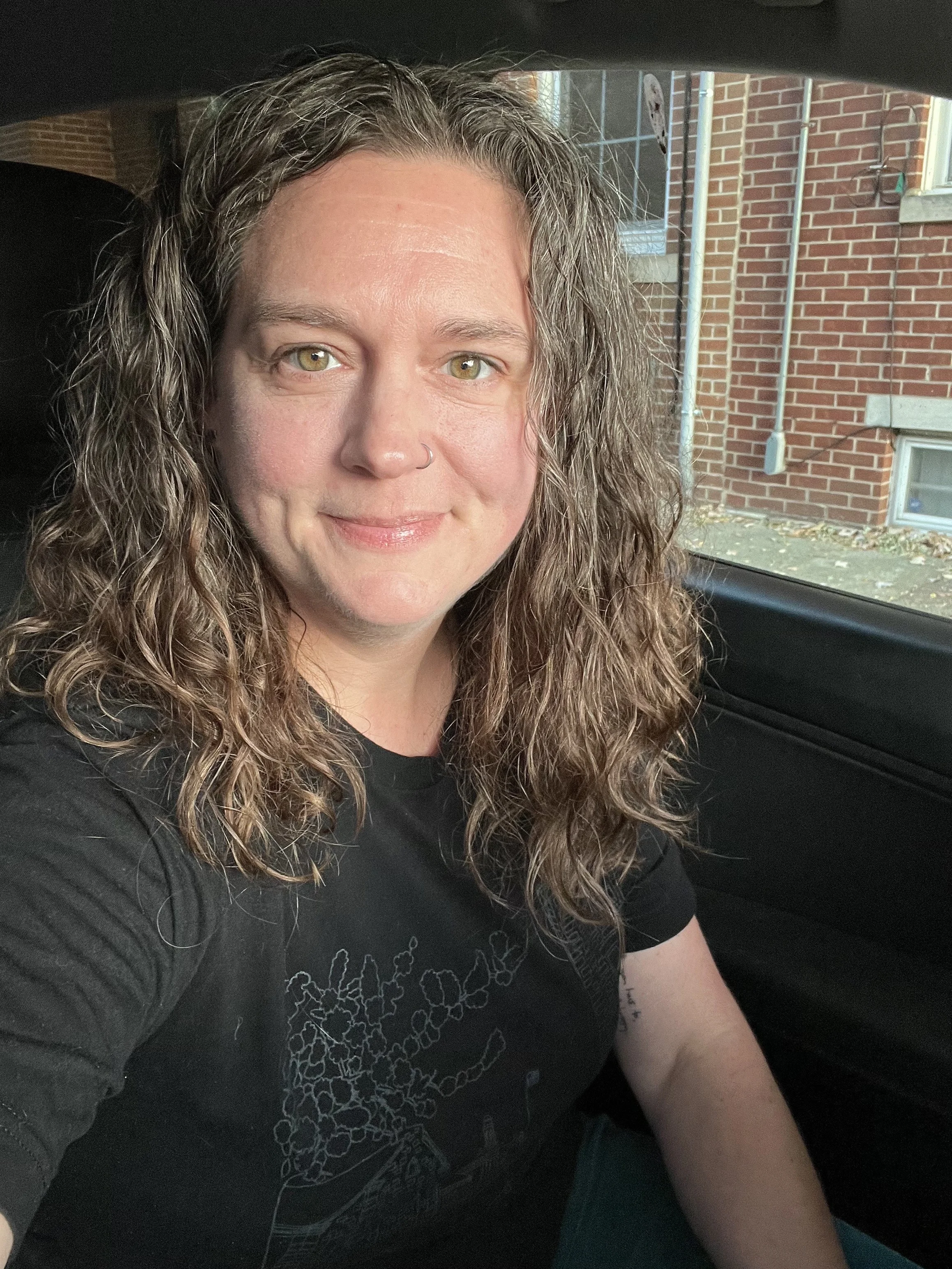 A woman with curly hair and a nose piercing smiling while sitting in a car, with a brick building visible outside the window.