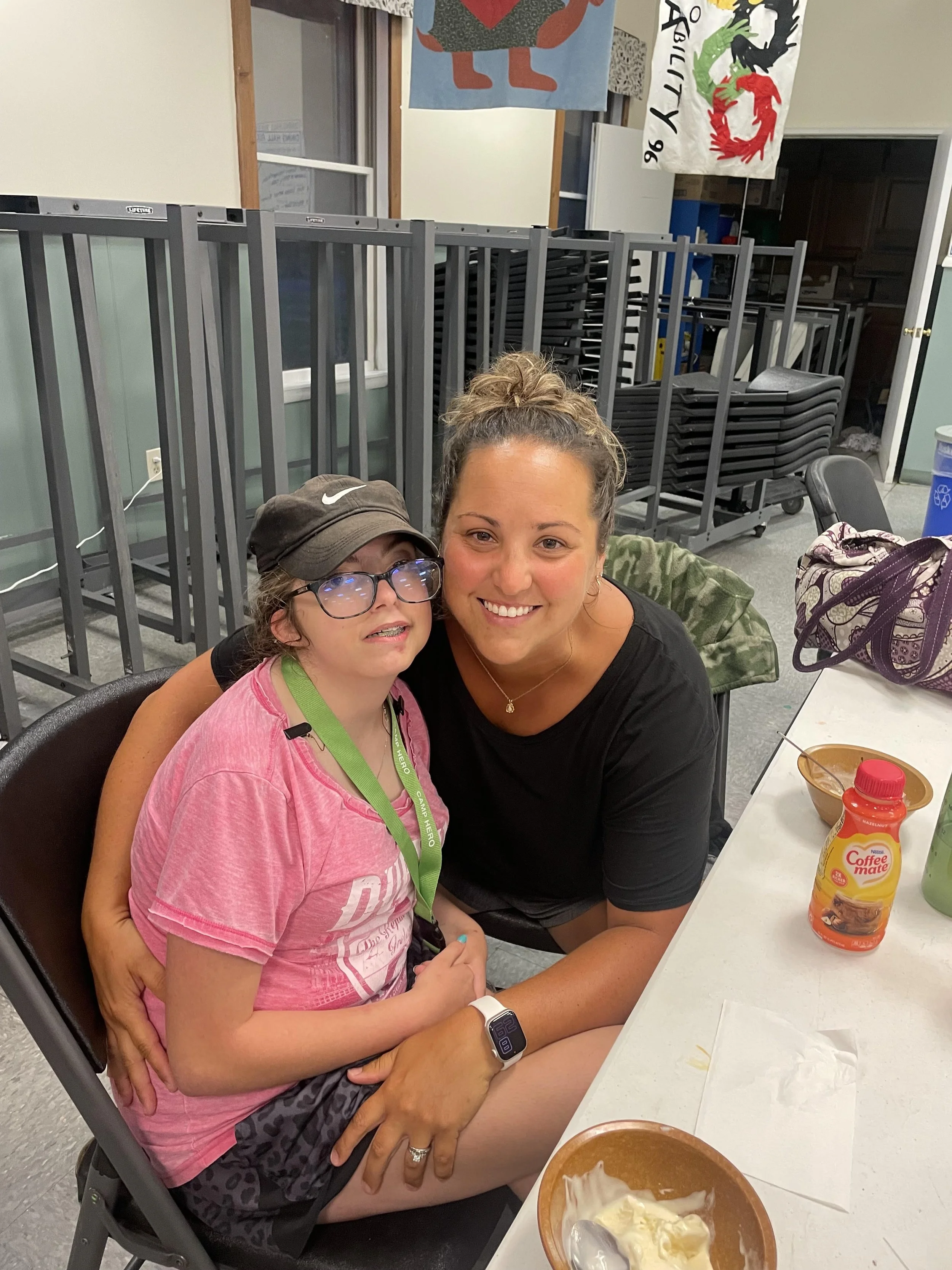 A woman and a girl sitting together at a table, smiling for the camera indoors. The girl is wearing glasses, a pink shirt, and a black cap, with a green lanyard around her neck. The woman is wearing a black shirt. On the table, there is a bowl of ice cream, a bottle of Coffee Mate creamer, a cup, and some other items.