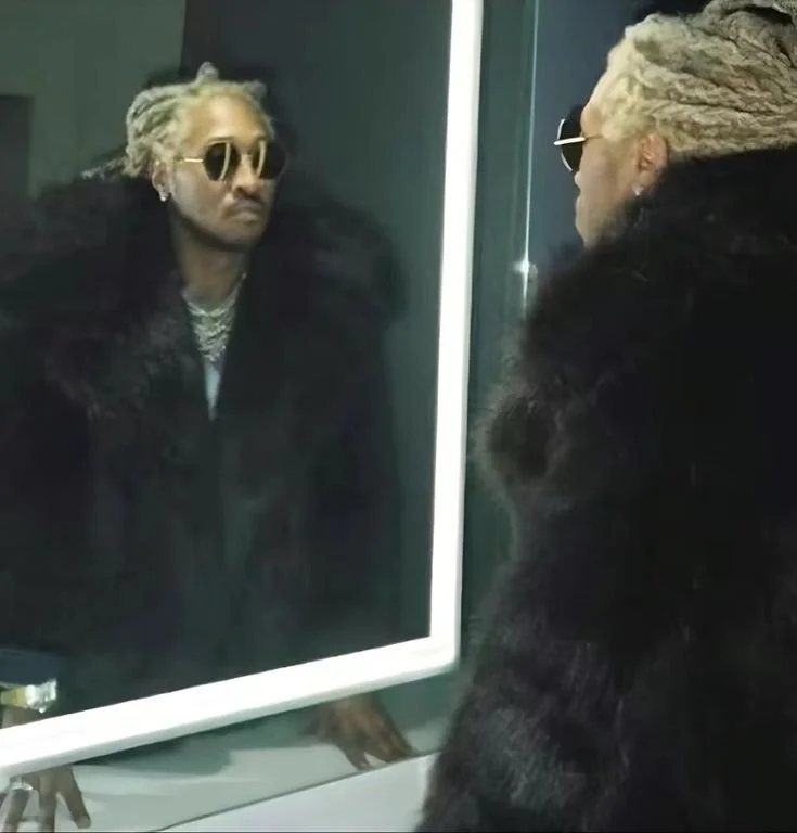 Person with blonde dreadlocks wearing sunglasses and a black fur coat looking at their reflection in a mirror.