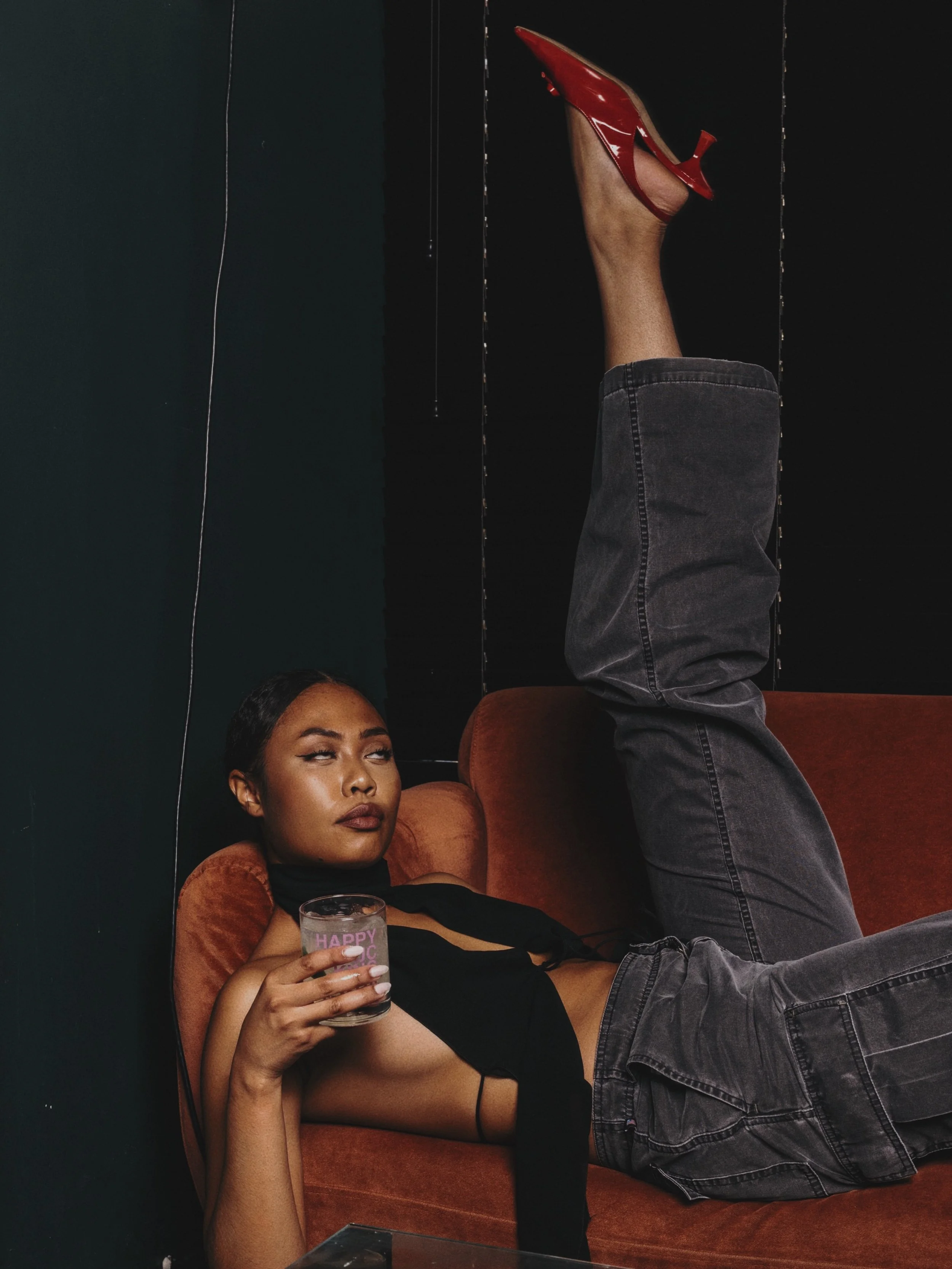 A young woman with dark hair, wearing a black top and grey jeans, lying on an orange velvet couch. She is holding a glass of drink in her right hand and looking contemplatively to the side. One of her legs is raised straight up, showing a red high-heeled shoe against a black background.