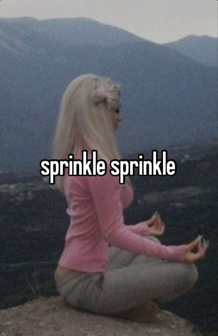 A woman with long blonde hair, dressed in a pink sweater and gray pants, sitting cross-legged in a meditative pose outdoors with mountains in the background. The text 'sprinkle sprinkle' is overlaid on the image.