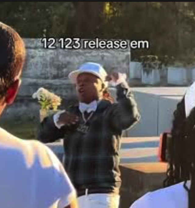 Man wearing a cap and a leather jacket speaking during an outdoor event with people and trees in the background.