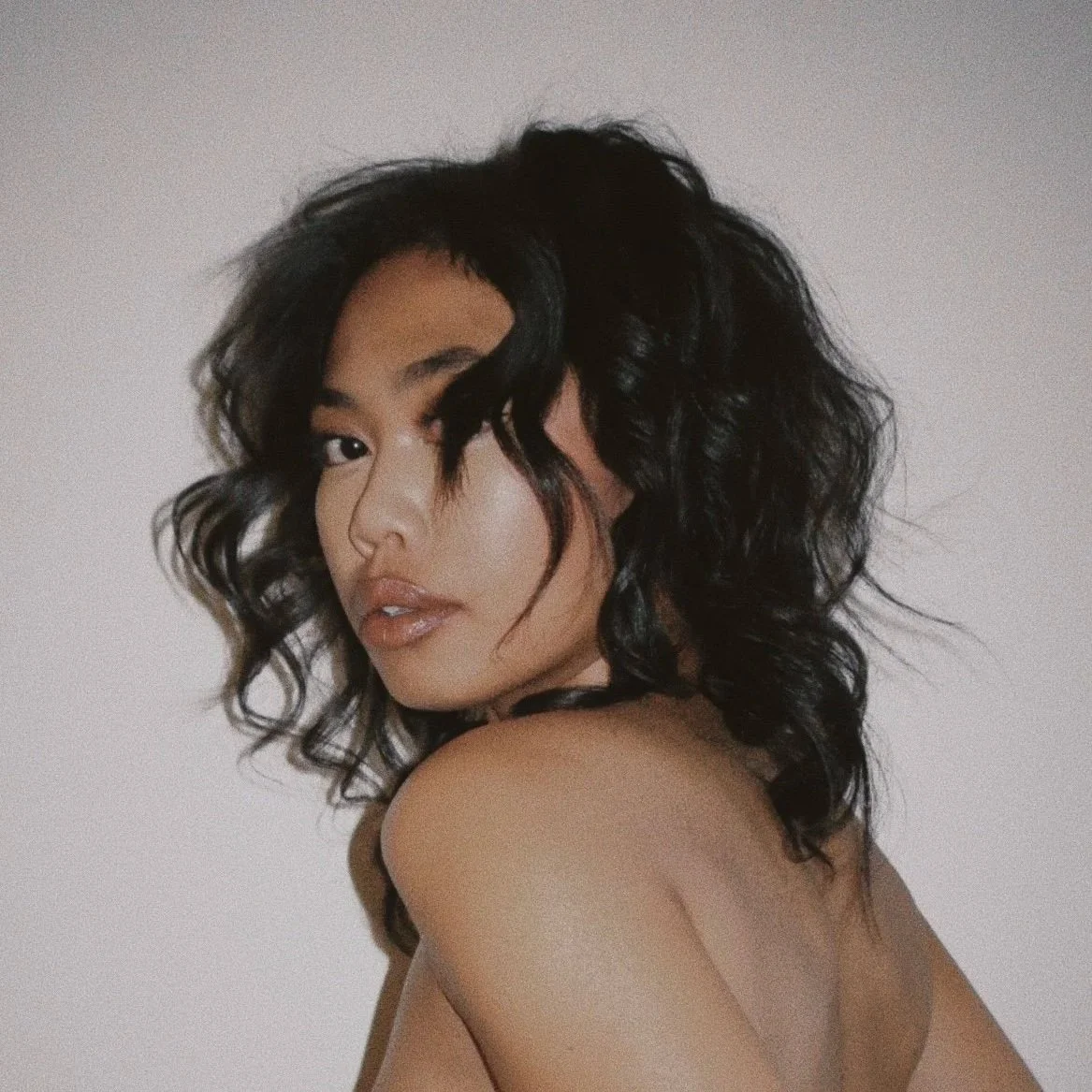 A woman with curly black hair looking over her shoulder with a neutral expression against a plain white background.