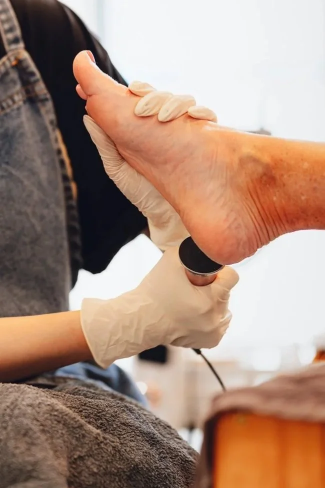 A healthcare professional in white gloves is examining an elderly person's foot using a handheld device in a medical setting.