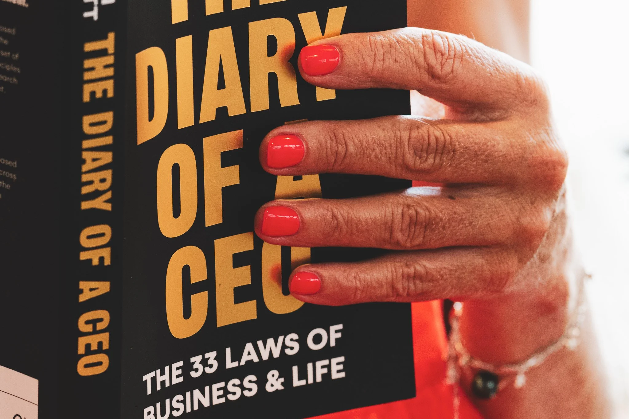 A hand with coral-colored nails holding a book titled 'The Diary of a CEO: The 33 Laws of Business & Life'