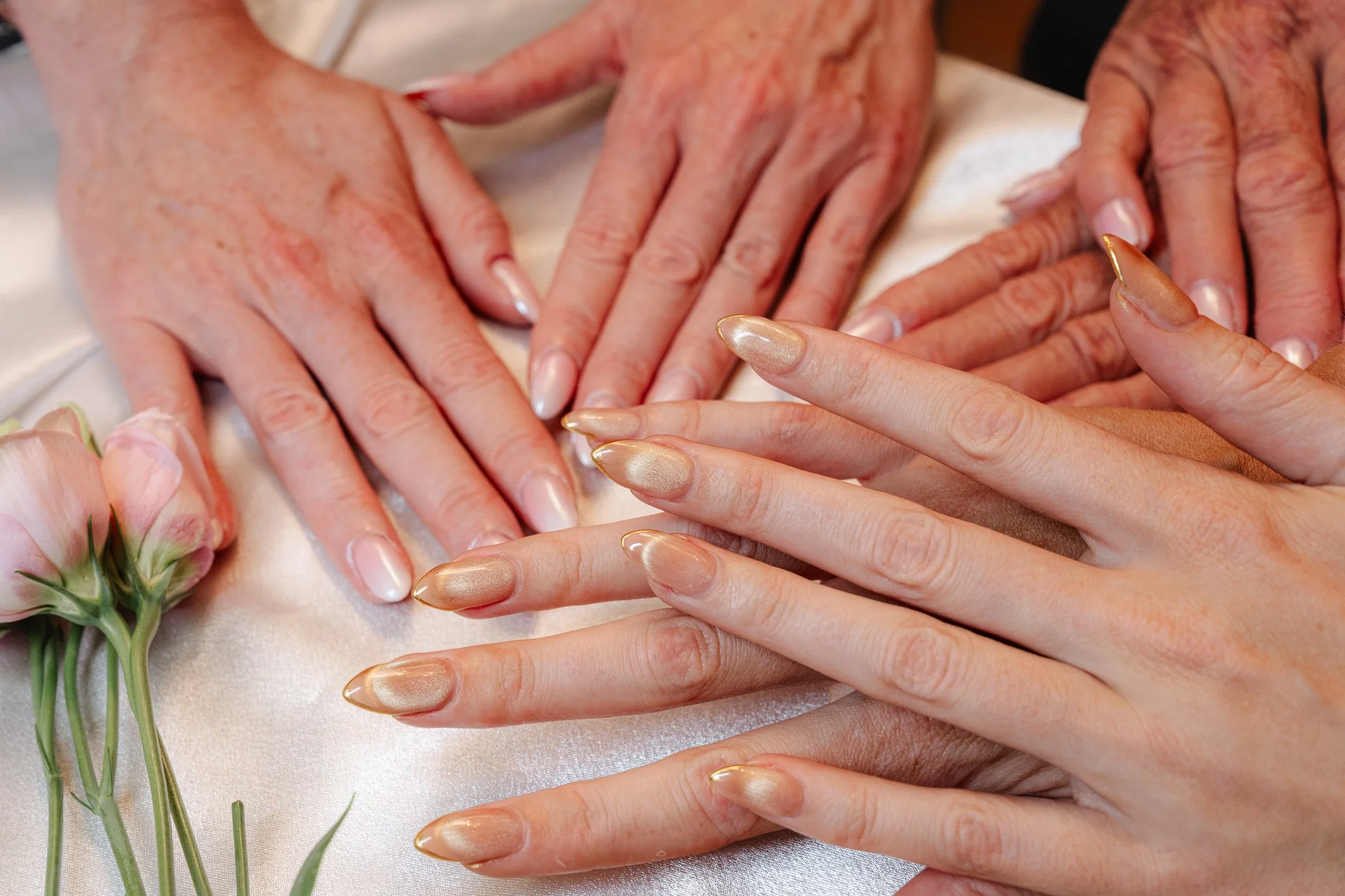 Close-up of several hands resting on a satin cloth, with some having manicured nails and some not, and a small bouquet of pink flowers nearby.