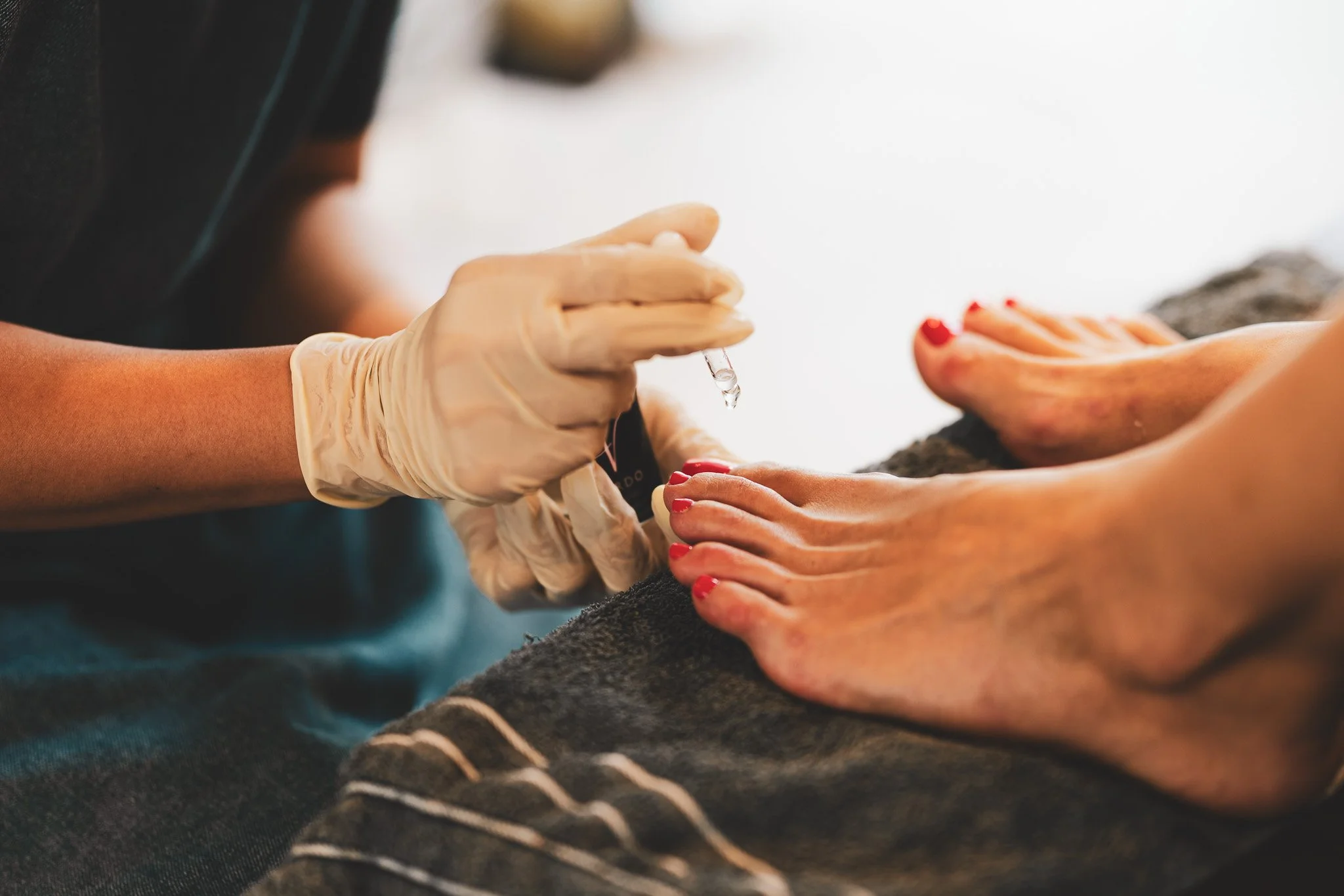 A person is getting a pedicure, with a worker wearing gloves applying clear polish to their toenails.